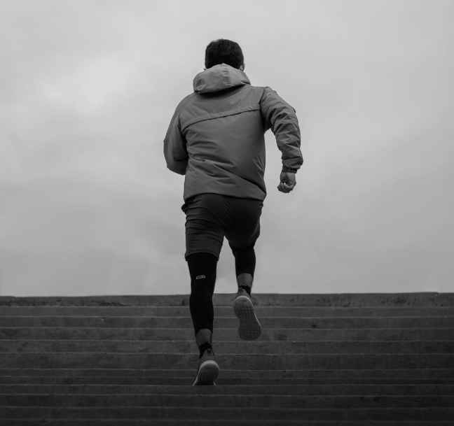 Person wearing a jacket and leggings running up concrete stairs against an overcast sky, shown from behind in black and white.