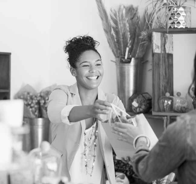 Smiling woman behind a counter handing a shopping bag to another person in a store with decorative plants and jars in the background.