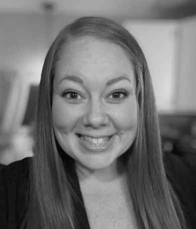 Black and white close-up portrait of a woman with long straight hair smiling at the camera in an indoor setting.