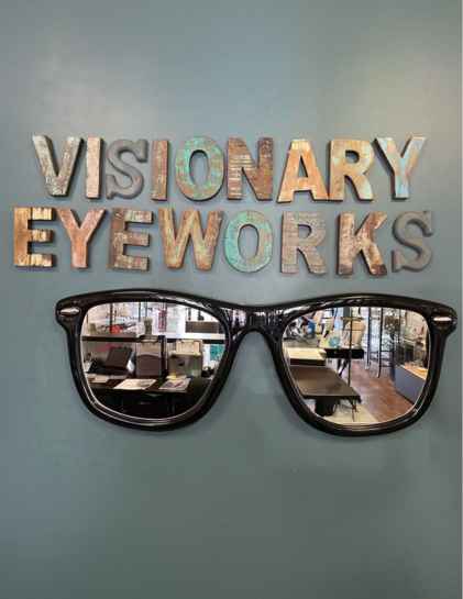 Decorative wall display with the words 'VISIONARY EYEWORKS' in textured, multi-colored letters above a large black eyeglass frame reflecting the interior of an eyewear store.