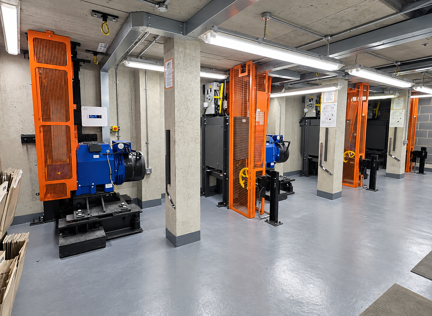 Industrial machinery with blue motors and orange safety guards arranged along concrete pillars in a clean, well-lit room with gray floors and exposed ceiling pipes.