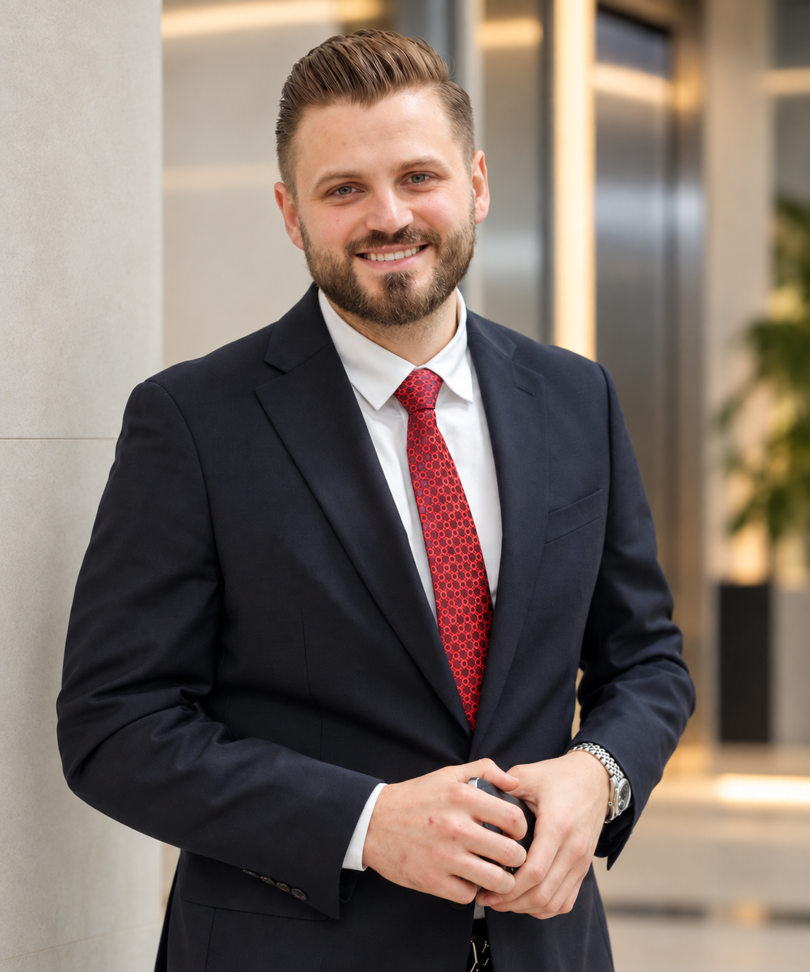 Smiling man with a beard wearing a dark suit, white shirt, and red patterned tie, standing indoors with blurred background and holding a small object in his hands.