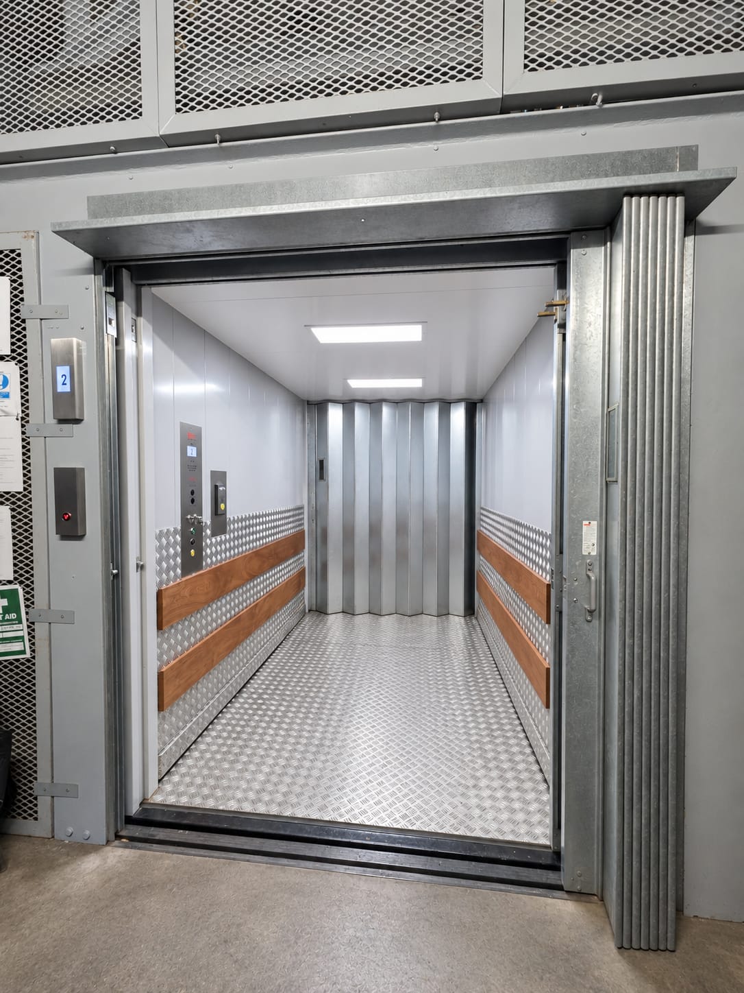 Open large industrial freight elevator with metal walls, diamond plate floor, wooden handrails, and accordion-style metal back door.