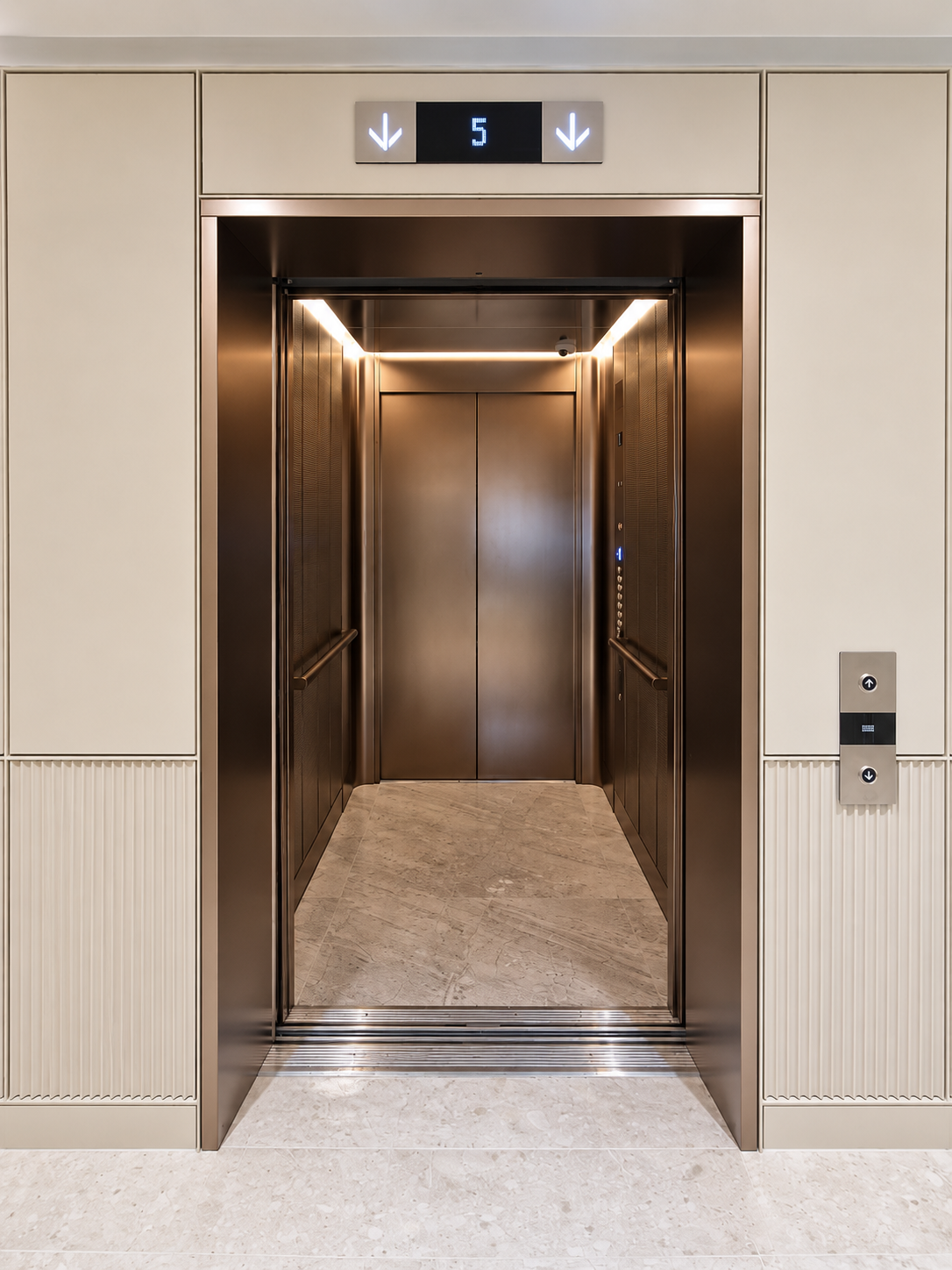 Open modern elevator with bronze interior and marble floor, floor indicator showing number 5 with two down arrows above the doors, and call buttons on the right wall.