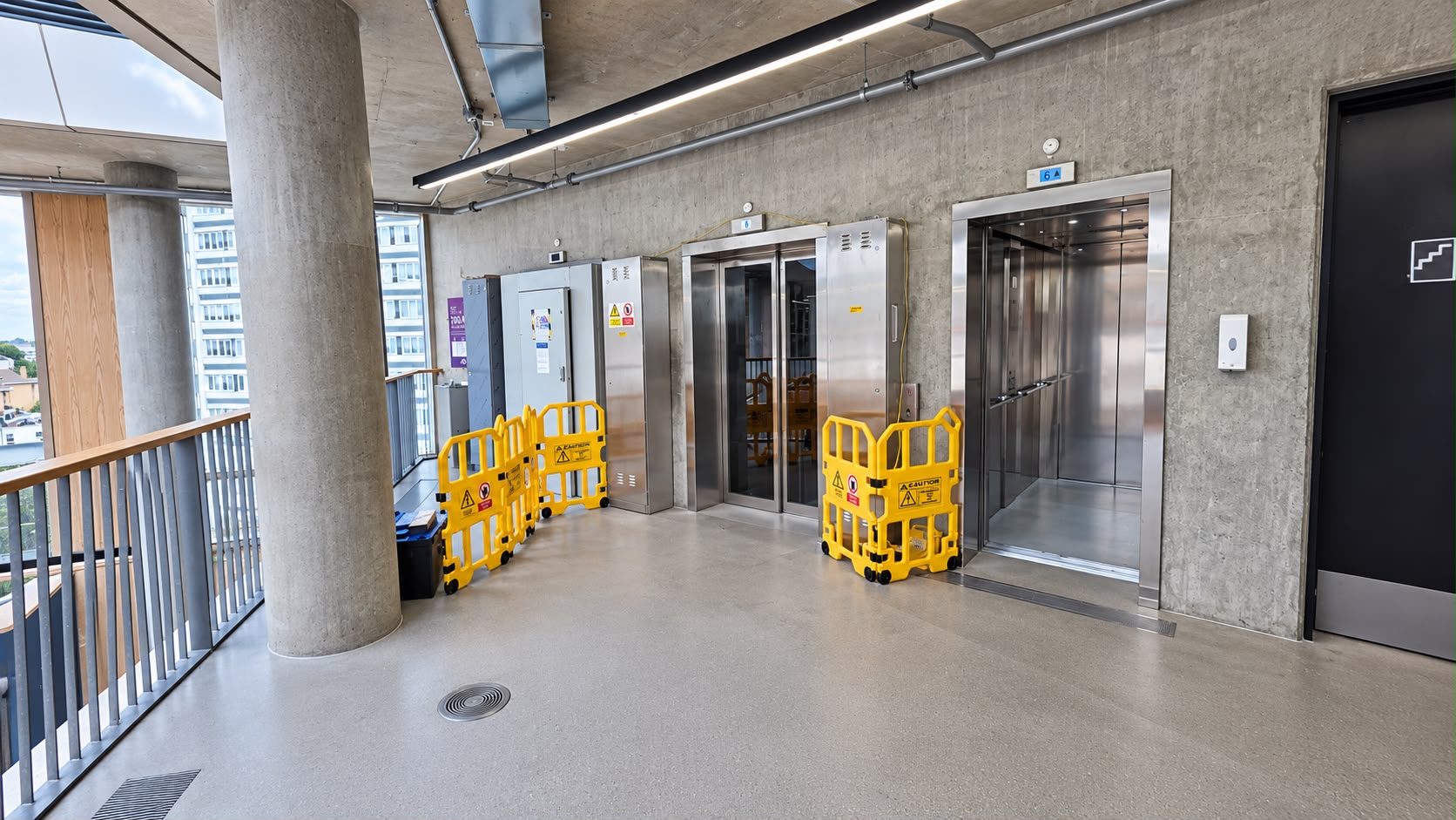 Modern building corridor with two stainless steel elevators, one with open doors, surrounded by yellow caution barriers, concrete walls and floor, large concrete pillar, metal railing, and a black door with a stair icon on the right.