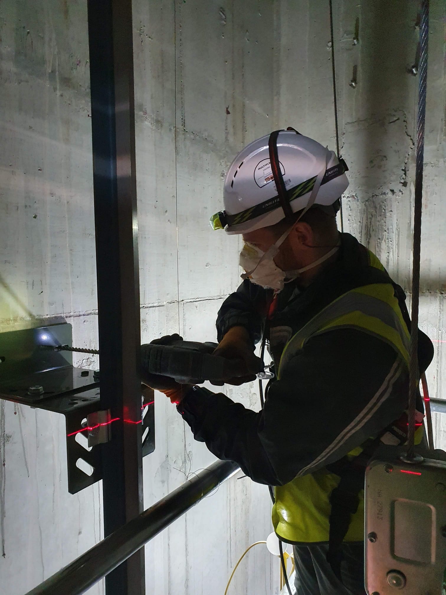 Construction worker wearing a white safety helmet with a headlamp, a face mask, and a yellow reflective vest, using a power drill to work on a metal structure mounted on a concrete wall, illuminated by a red laser level line.