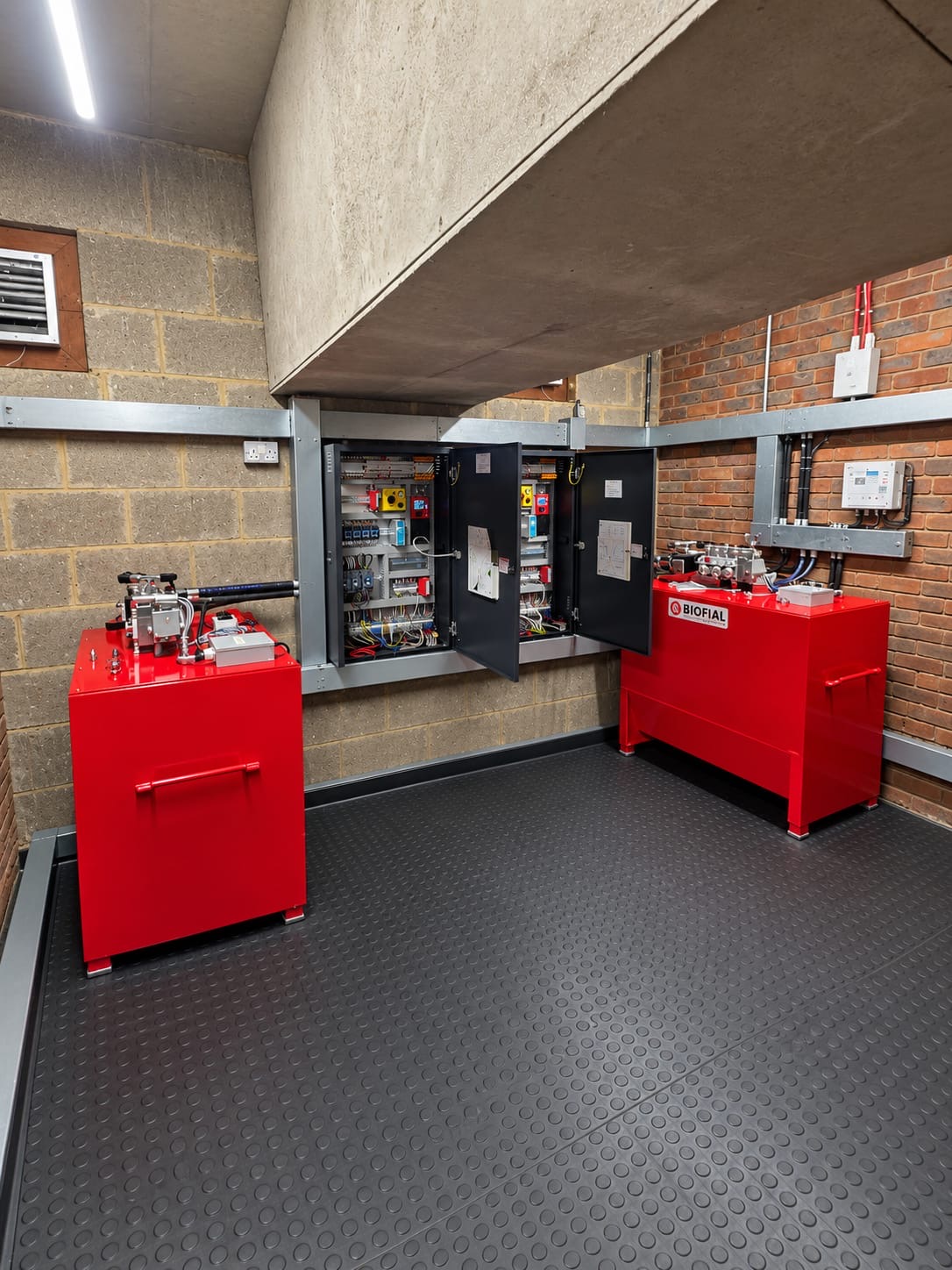 Industrial room with brick and concrete walls housing two red metal machinery units labeled Biofial, positioned on a black rubberized floor with circular patterns, and open electrical control panels mounted on the wall between them.