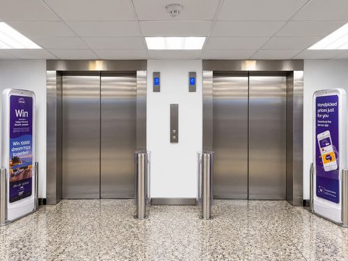 Two closed stainless steel elevator doors in a clean, modern lobby with terrazzo flooring, white walls, and ceiling lights; digital floor indicators display '0' above each elevator, with two vertical cylindrical stainless steel bollards between them and advertising signs on either side promoting the Nectar app and a prize draw for dream trips.
