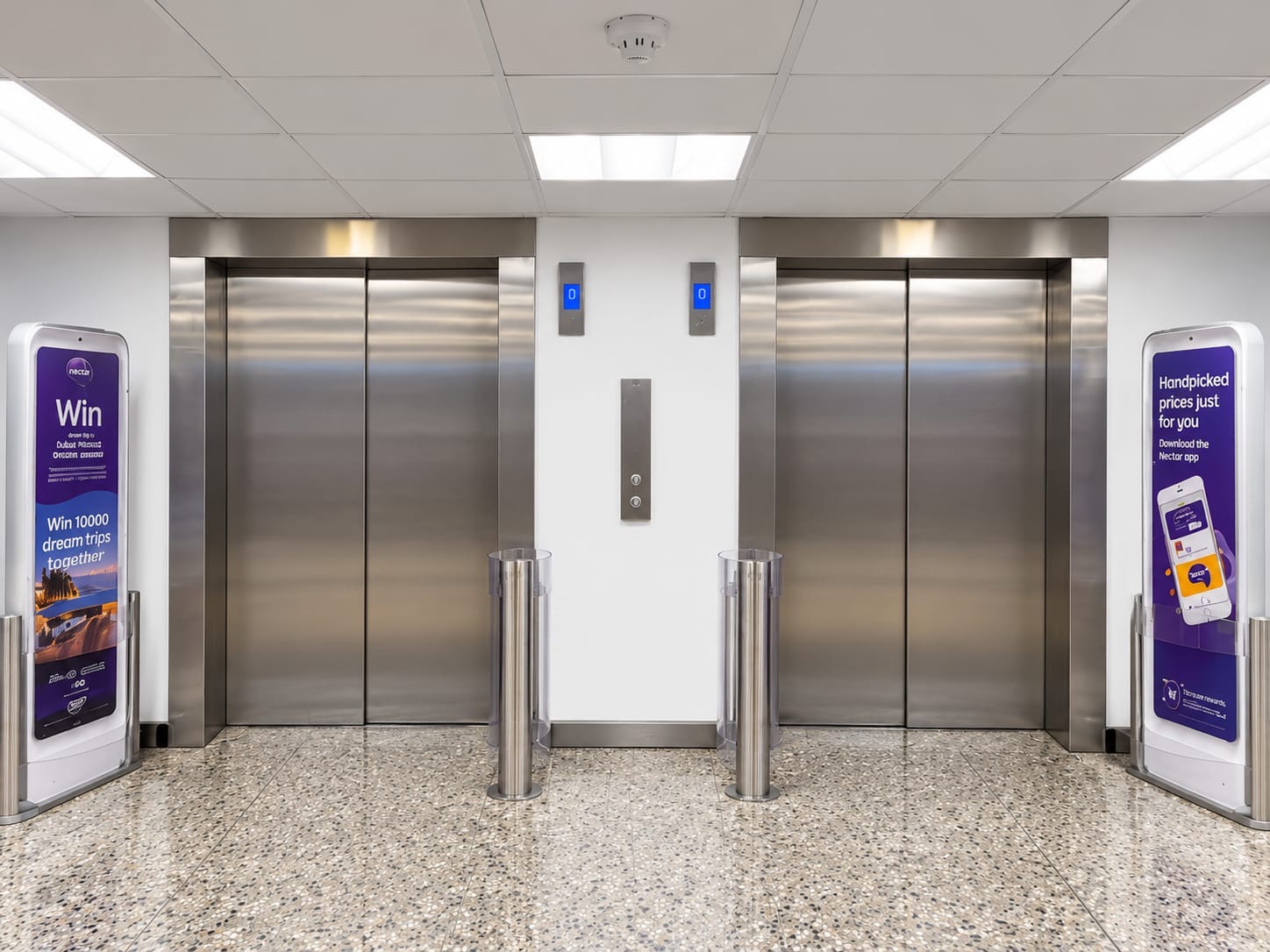 Two closed stainless steel elevator doors in a clean, modern lobby with terrazzo flooring, white walls, and ceiling lights; digital floor indicators display '0' above each elevator, with two vertical cylindrical stainless steel bollards between them and advertising signs on either side promoting the Nectar app and a prize draw for dream trips.