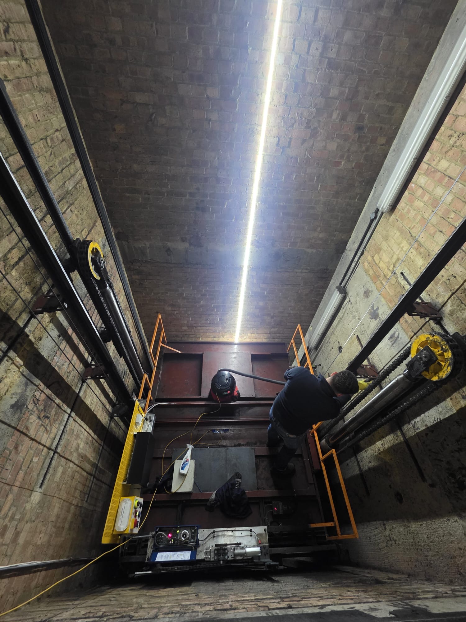 Man working inside an elevator shaft with brick walls, engine machinery, and yellow safety railings.