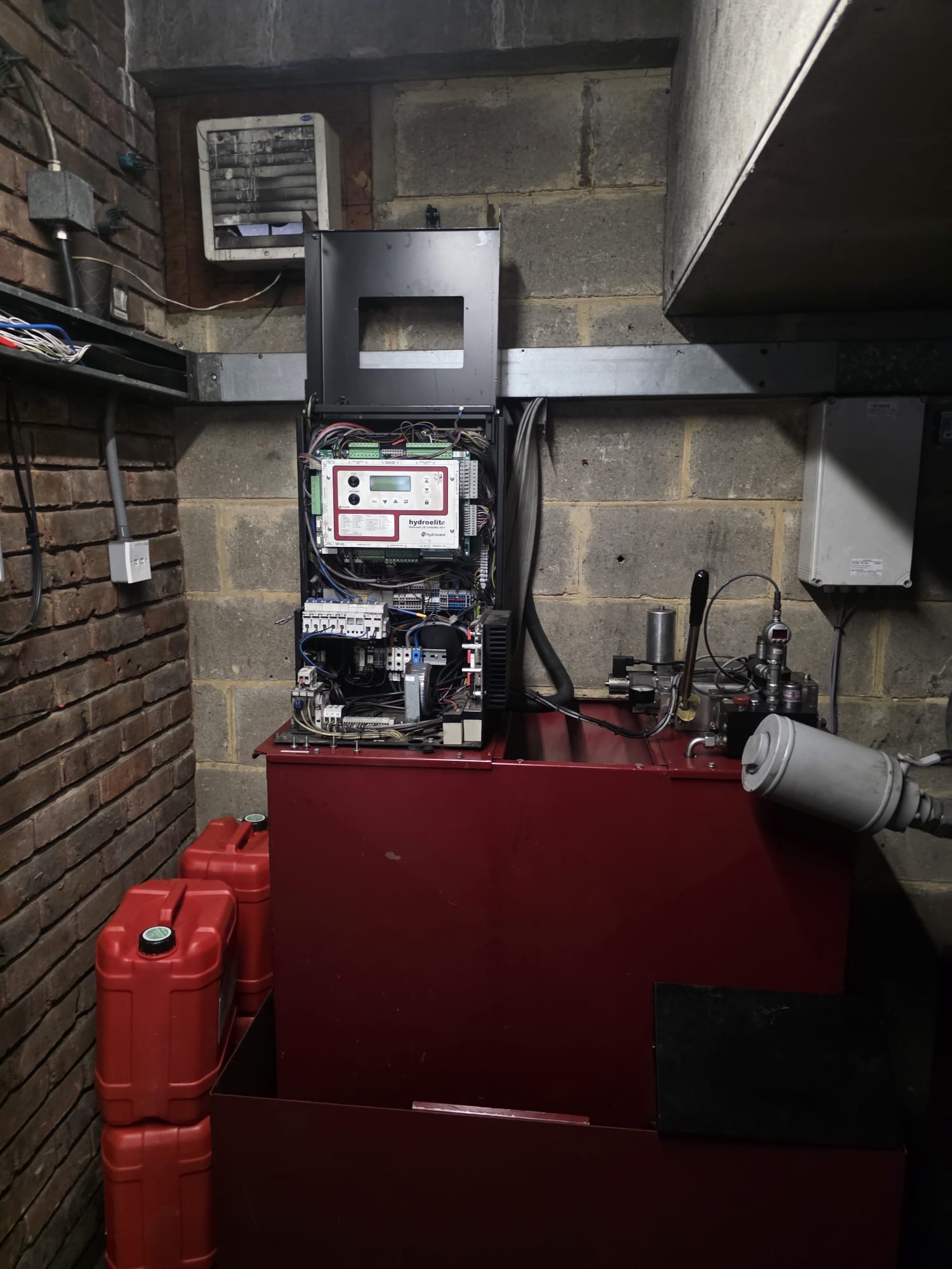 Industrial control panel with exposed wiring and a display labeled Hydroelite mounted on a large red metal unit against a concrete block wall, with red plastic containers stacked beside it.