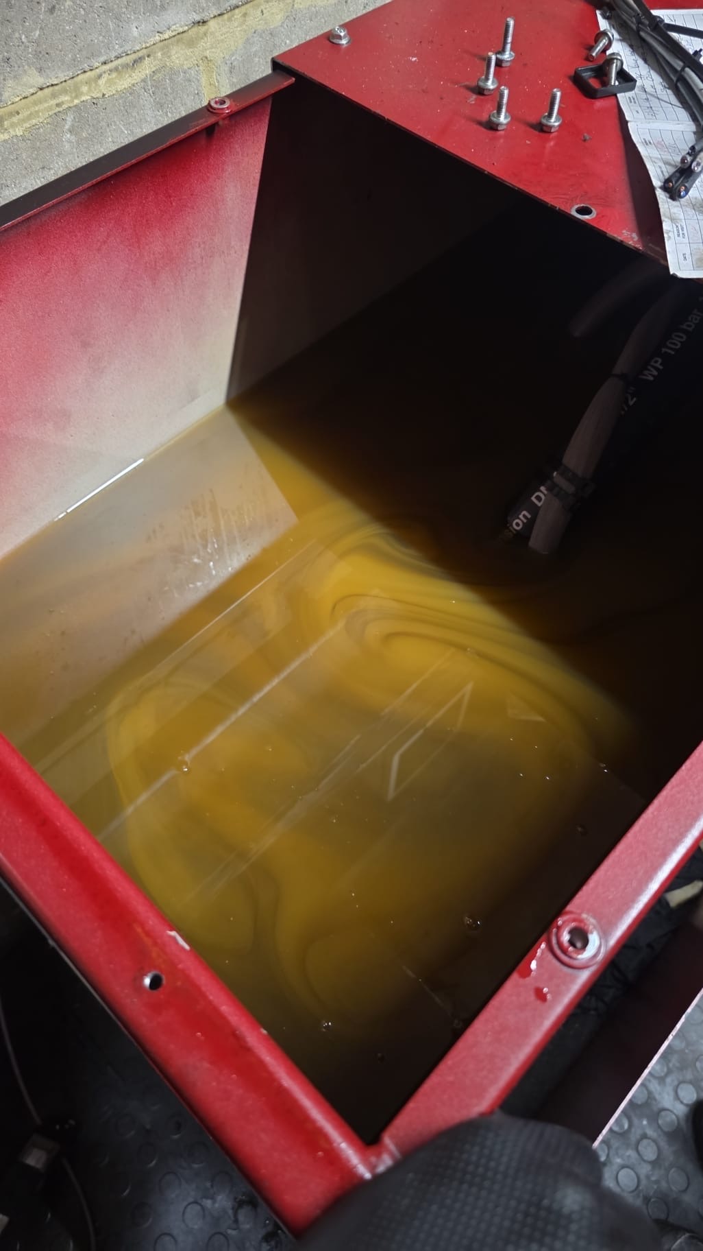 Close-up of a red metal container partially filled with yellowish liquid, with several metal bolts lying on the container's edge and some black hoses or cables nearby.