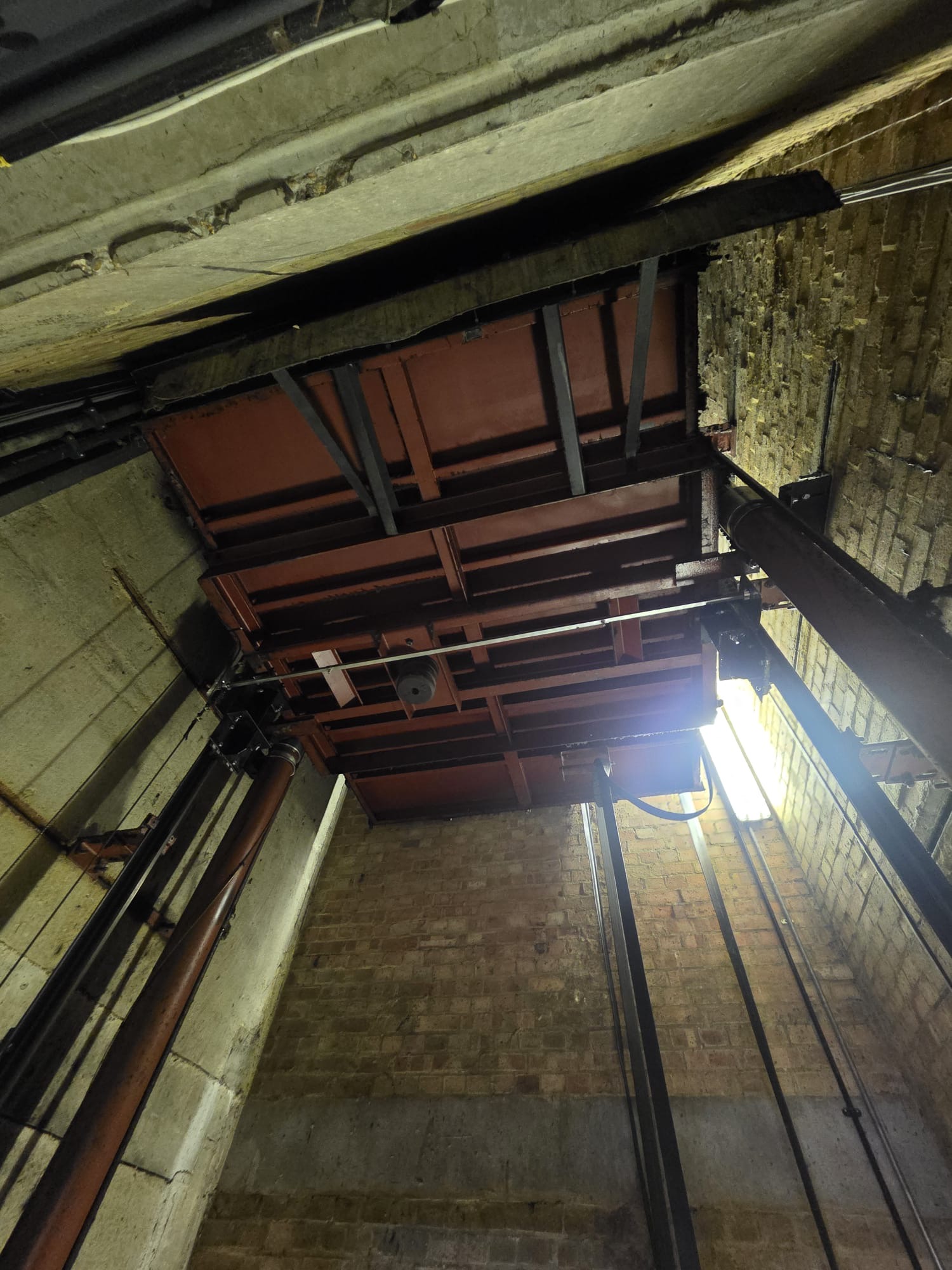 View looking up inside an industrial shaft or elevator pit with metal beams, pipes, brick and concrete walls, and a bright light on one side.