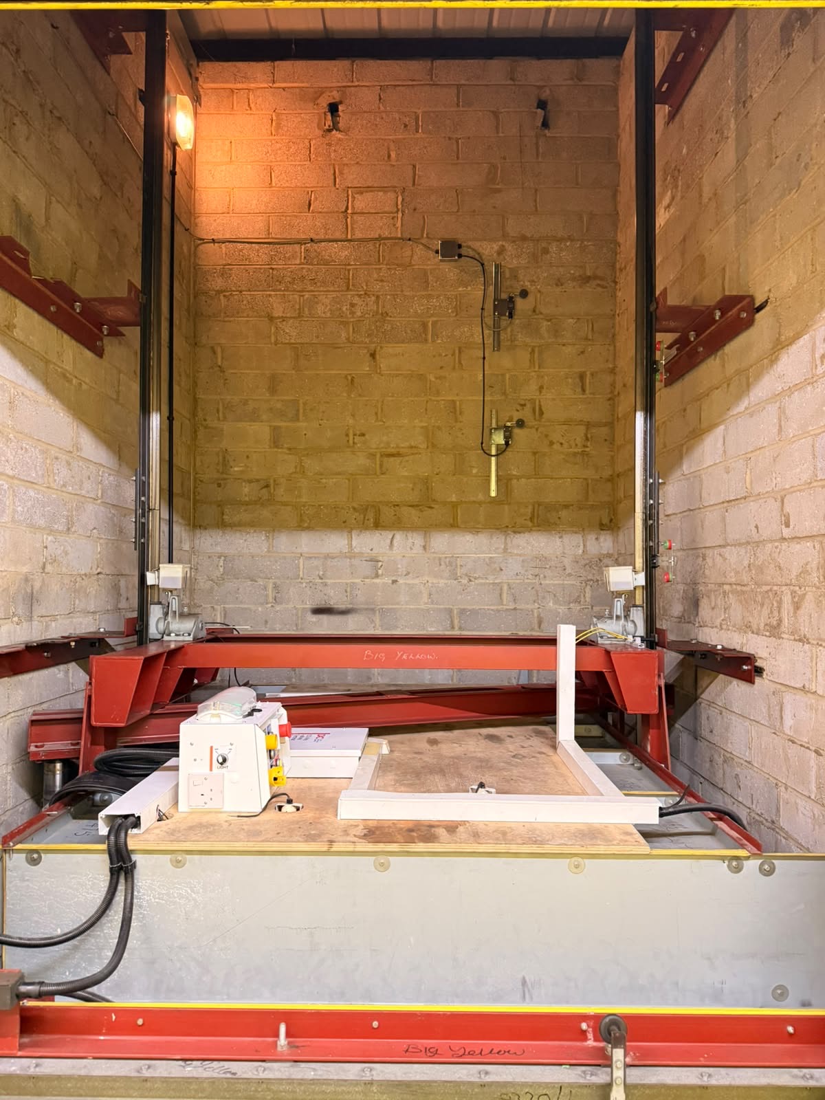Industrial mechanical equipment setup inside a brick-walled room with red metal framework, wooden platform, electrical components, and cables.