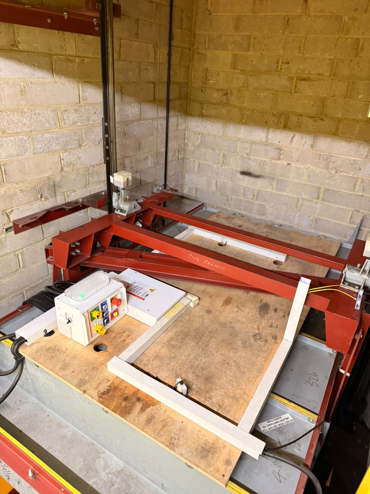 Mechanical assembly featuring a wooden platform with red metal supports inside a concrete block-walled room, equipped with an electrical control box and yellow wiring.