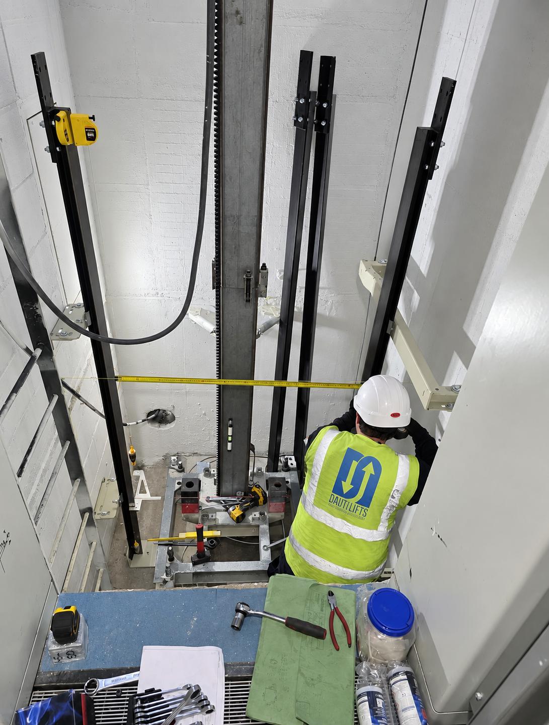 Worker in a white hard hat and yellow high-visibility vest with Dautlifts logo inspects or assembles components inside an elevator shaft with metal framework, tools, and measuring tape.