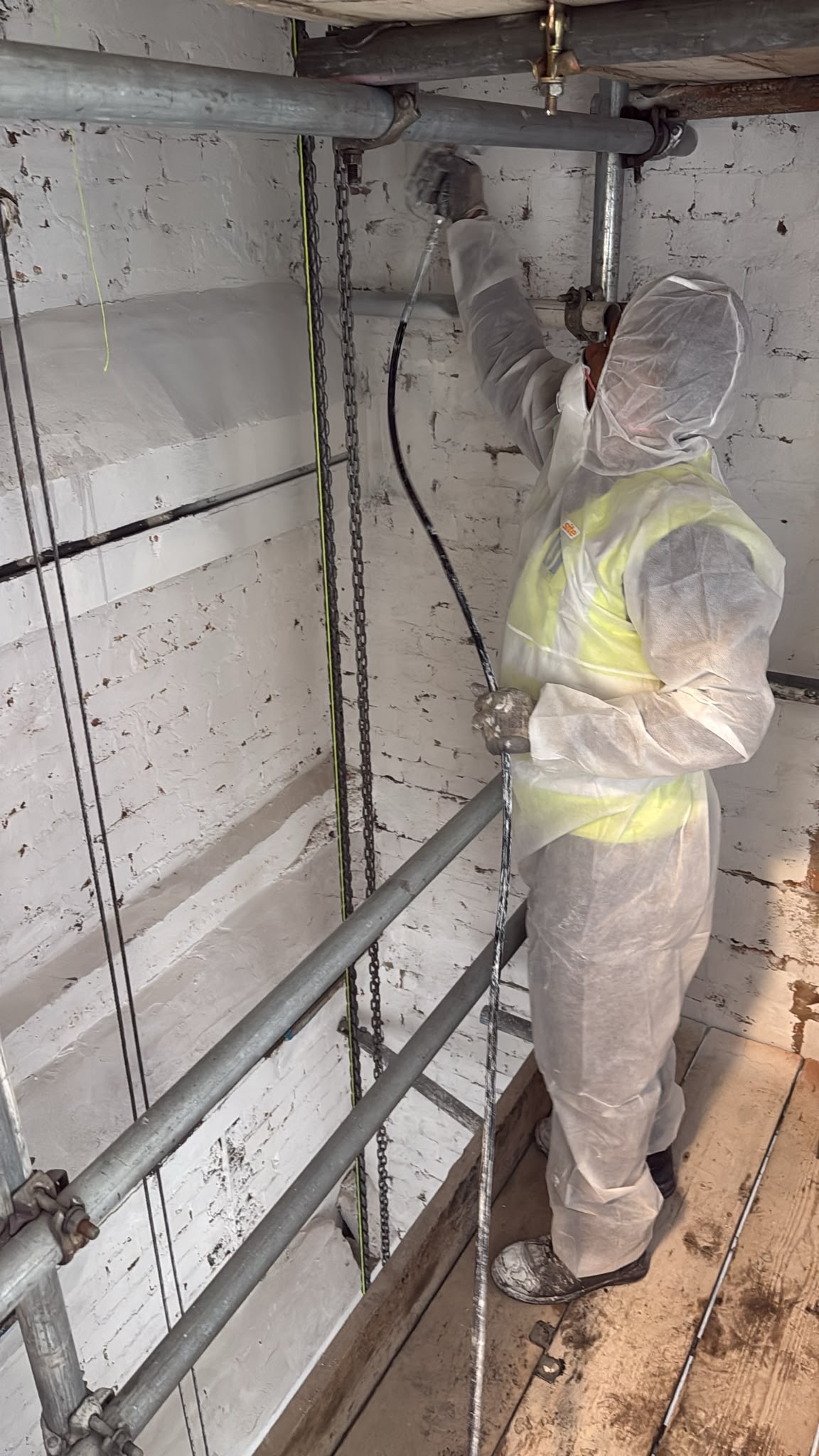 Worker in a white protective suit, gloves, and mask using a tool to polish or grind metal pipes in an industrial setting with white painted brick walls and metal scaffolding.