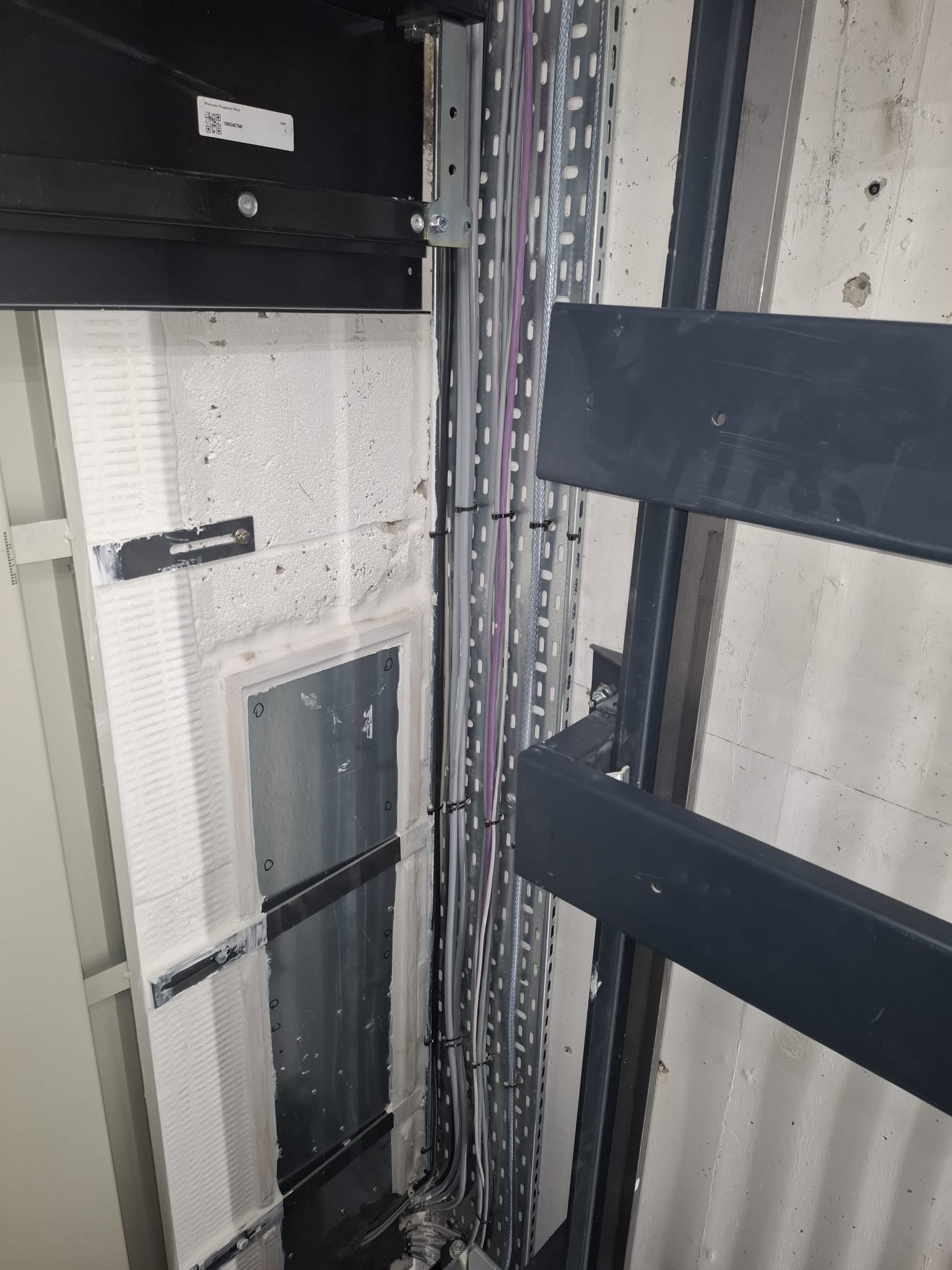 Corner of a utility room wall with electrical conduits and cables secured by zip ties running vertically within a metal perforated cable tray, adjacent to metal brackets and a partially visible black panel above.