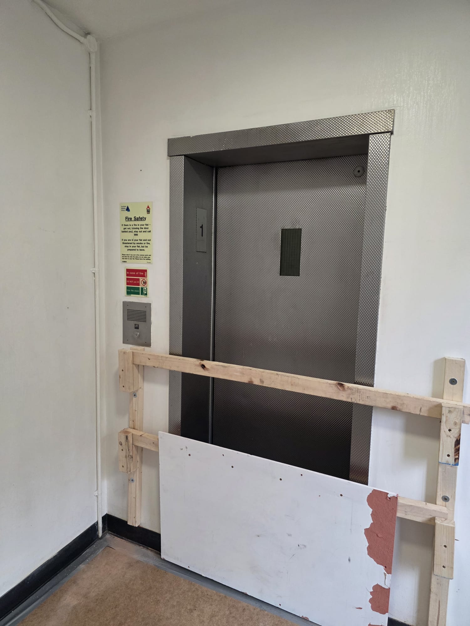 Closed metallic elevator door with the number 1 above the call button, surrounded by a temporary wooden and white board barrier, inside a plain white-walled hallway.