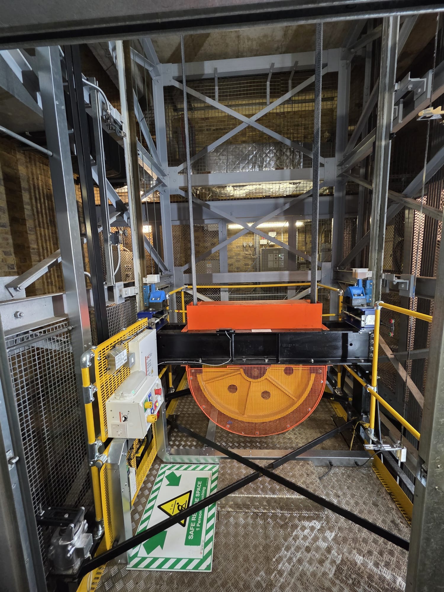 Interior of an elevator shaft featuring a large yellow and orange pulley wheel, steel cables, and safety rails with a sign on the floor indicating a safe refuge space for one person in case of a malfunction.