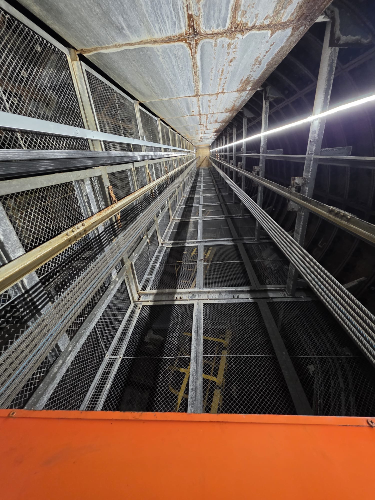 Looking upward inside an industrial elevator shaft showing metal framework, steel cables, mesh panels, and a rusty ceiling with a central light running along one side.