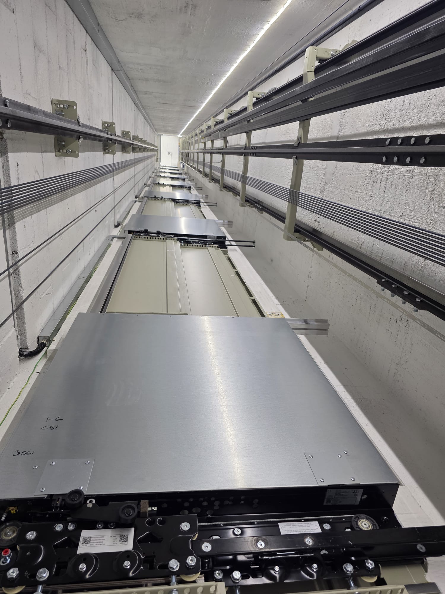 Inside view of an elevator shaft showing mechanical components and cables along the walls and ceiling in a concrete enclosure.