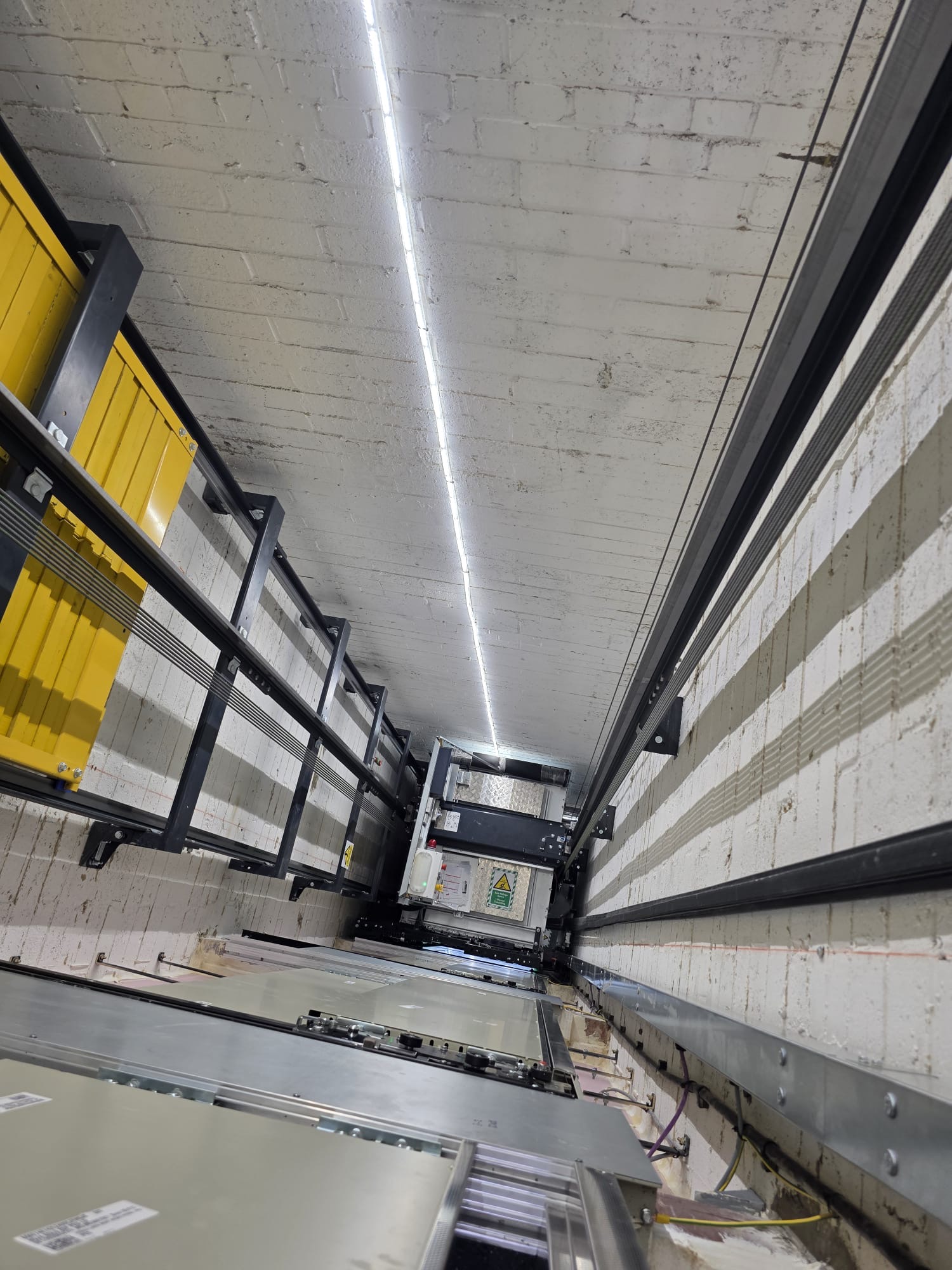Interior view of an elevator shaft showing metal rails, cables, and structural components with a white brick wall and a strip of LED lighting along the ceiling.