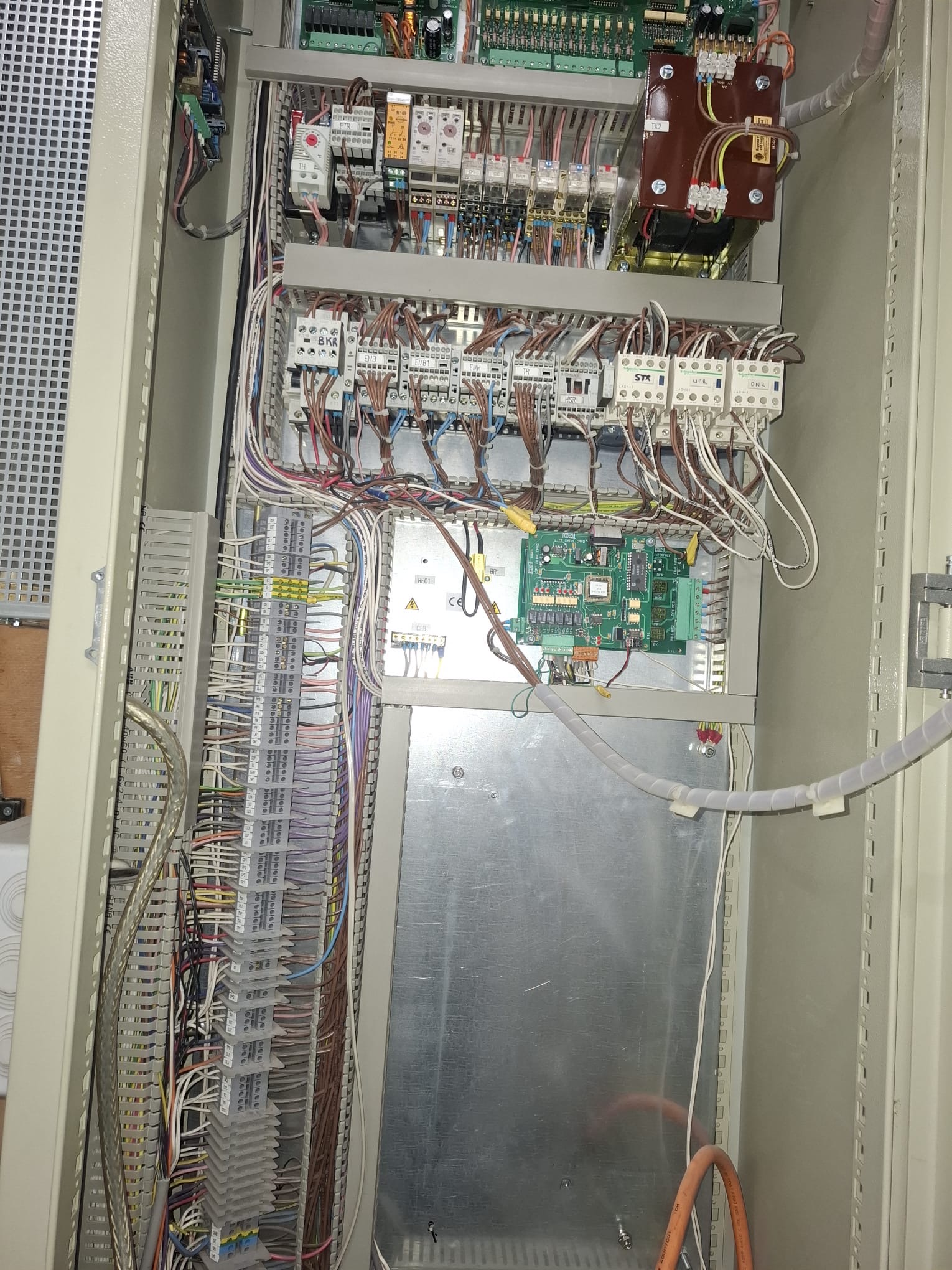 Open electrical control panel with multiple interconnected wires, circuit breakers, relays, and a green circuit board mounted inside a beige metal enclosure.