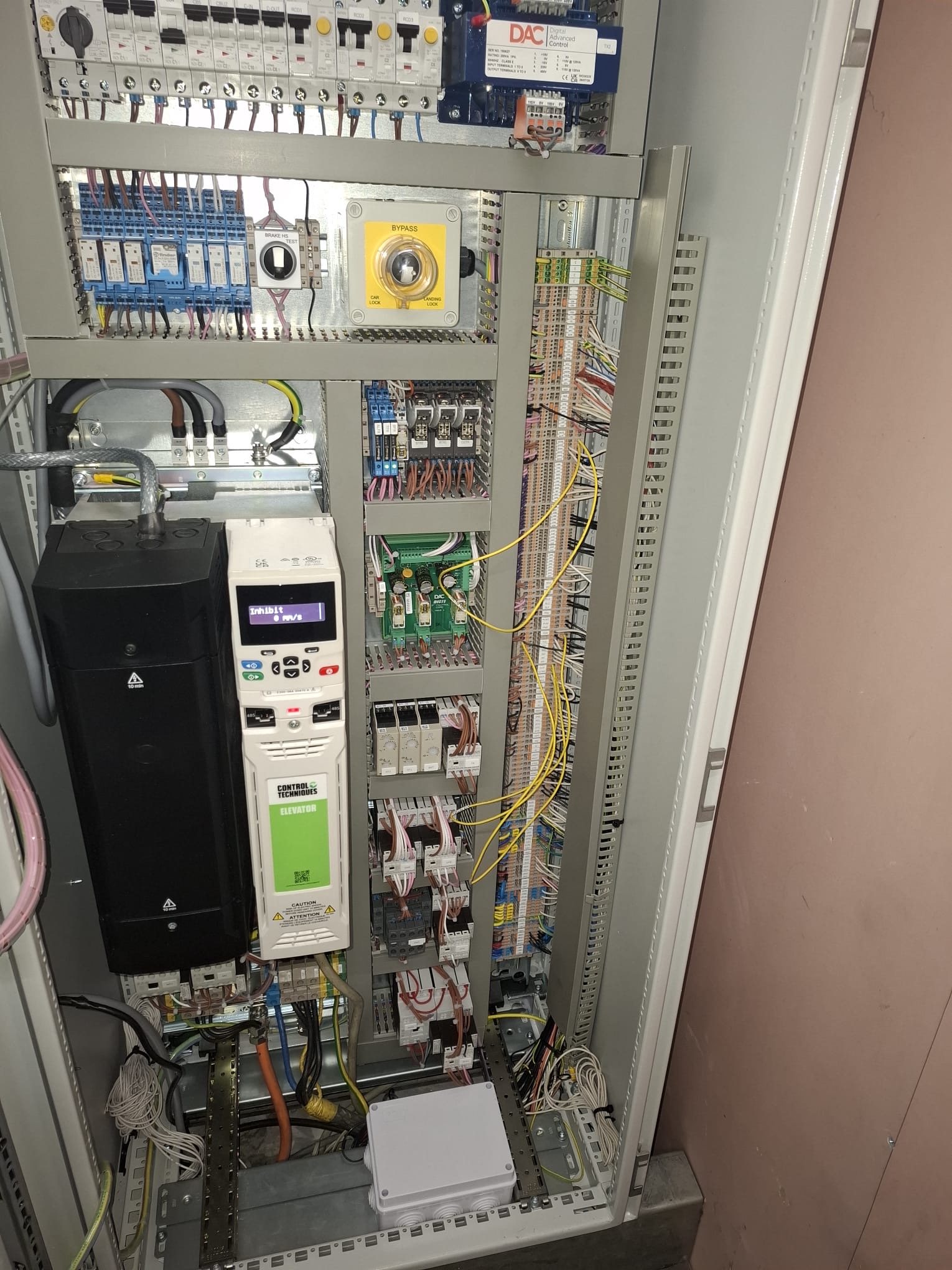 Open electrical control panel with a variety of circuit breakers, relays, wires, and a digital variable frequency drive labeled 'Control Techniques Elevator' showing a display with the word 'Inhibit'. The panel includes a yellow bypass switch and multiple organized wiring terminals.