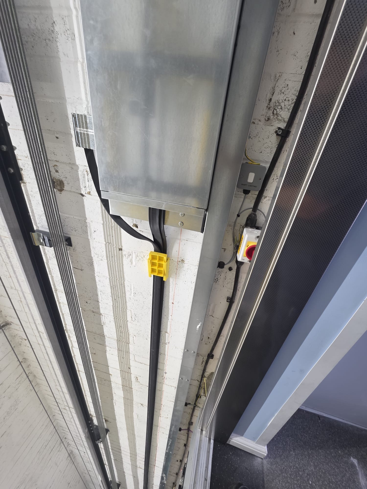 Ceiling view of a white brick wall with metal conduits and black cables running vertically alongside a large metal electrical box with a yellow clamp, a red emergency stop button, and a smart lighting switch panel.