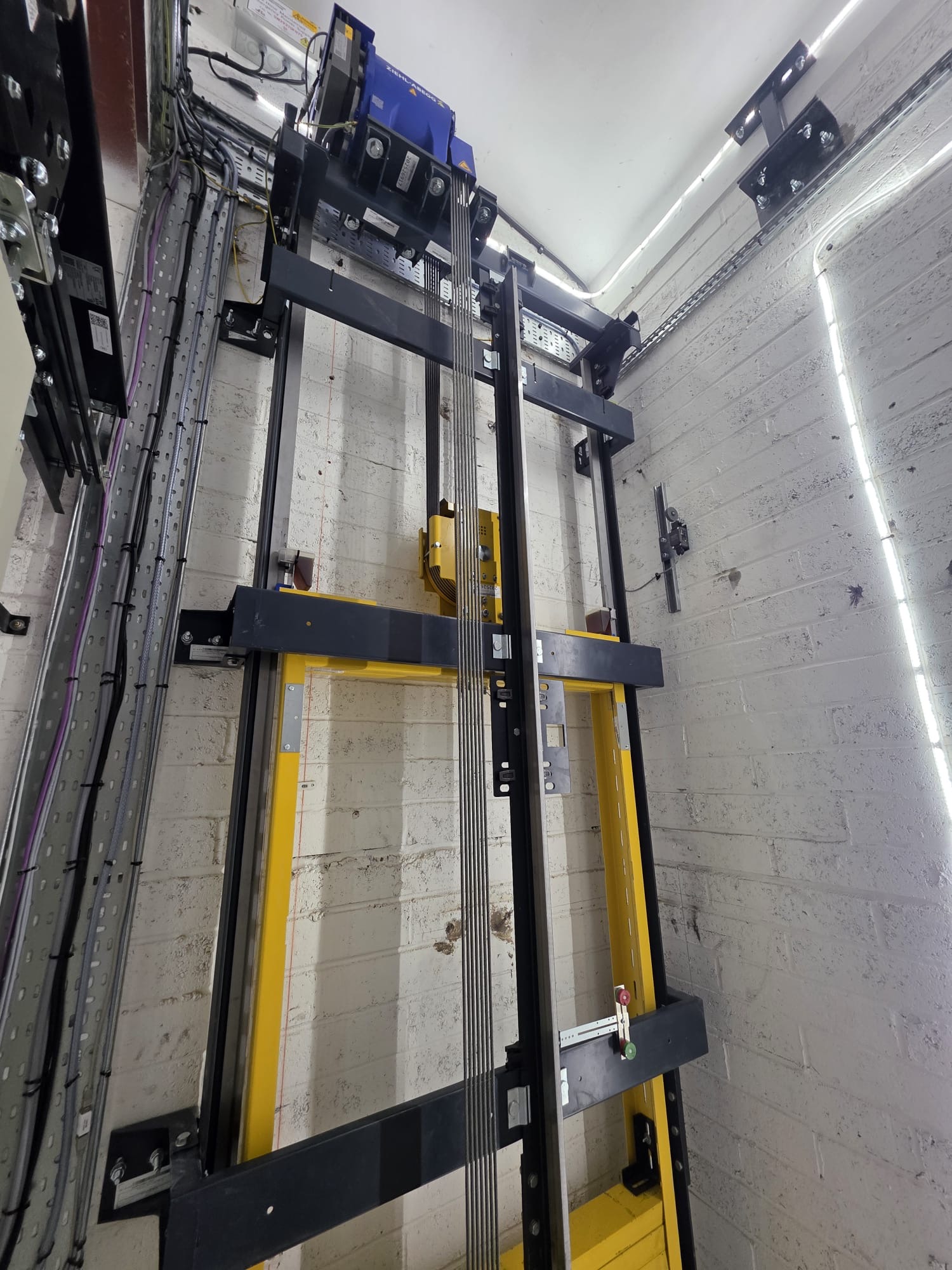 View looking upward inside an elevator shaft showing the yellow and black metal framework, steel cables, and machinery mounted at the top on a white brick wall background.