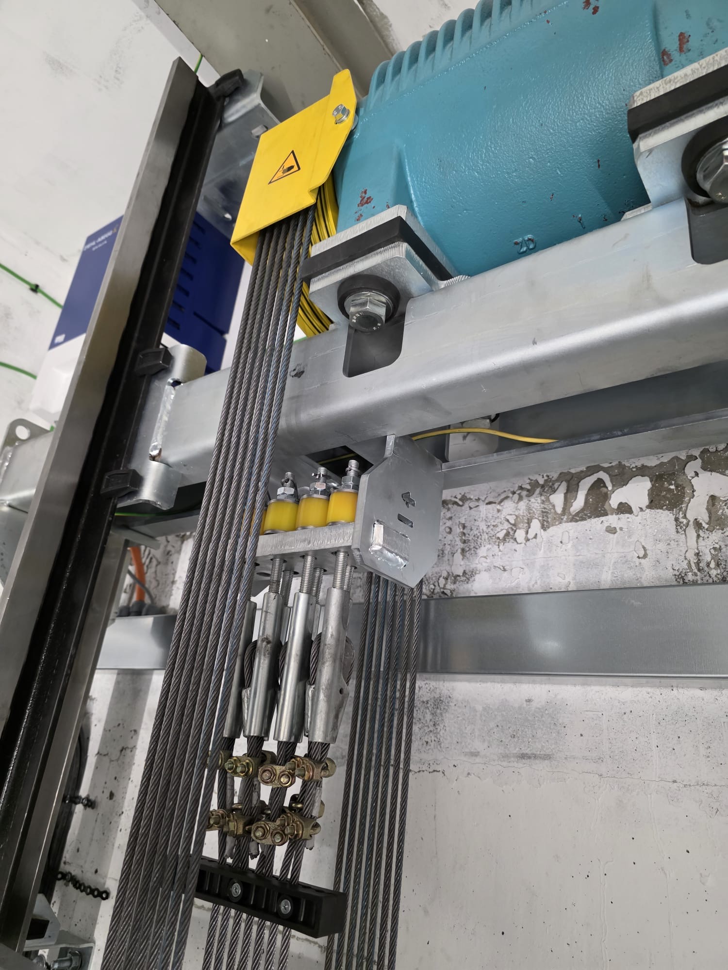 Close-up of an industrial mechanical assembly featuring thick steel cables attached to a metal frame with bolts, a yellow plastic component, and a blue motor unit in the background mounted on a wall with peeling paint.