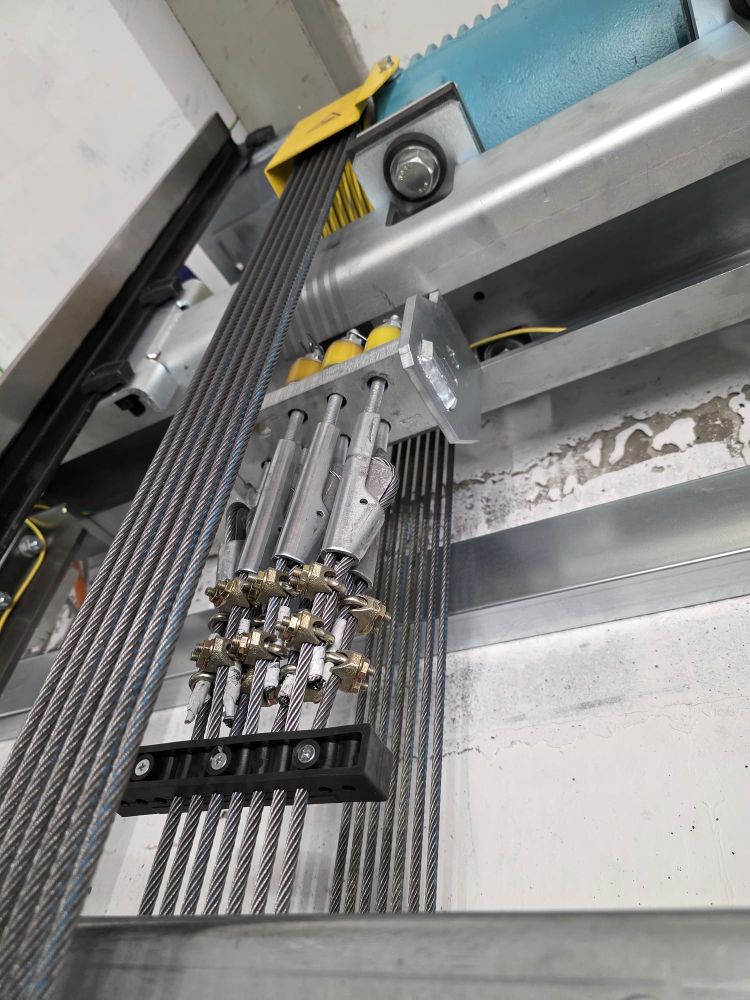 Close-up of multiple steel elevator cables secured with clamps, passing over yellow pulleys mounted on a metal frame inside an industrial setting with a concrete wall background.