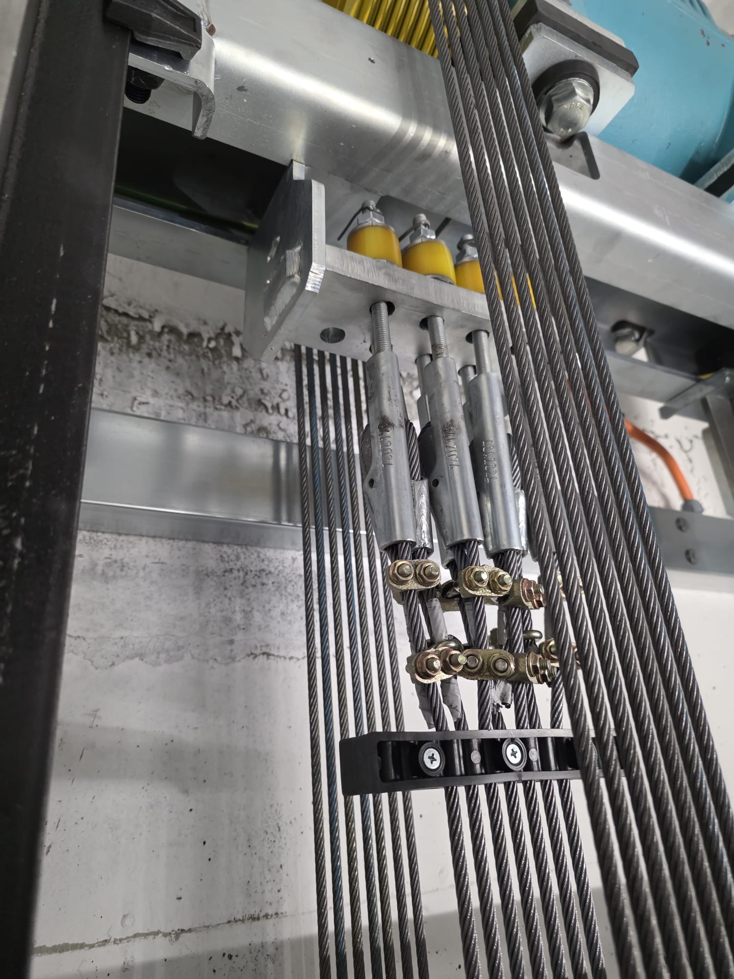 Close-up of multiple steel cables secured with metal clamps and threaded rods attached to a metallic support structure, with a white textured wall background.