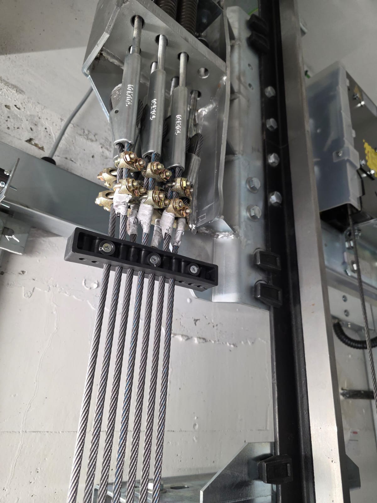 Close-up of six steel cables fastened with metal clamps and secured at the top with metal turnbuckles, attached to a metal frame mounted on a concrete wall.