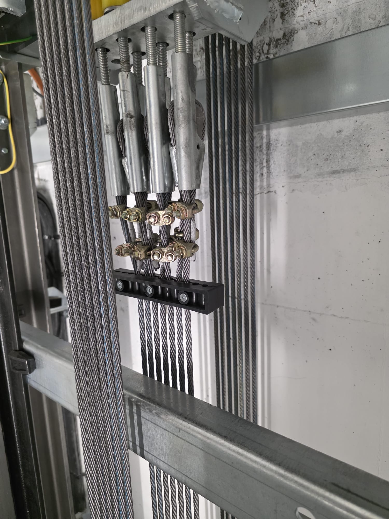 Close-up view of multiple tightly wound steel elevator cables secured by metal cable clamps and connected to metal brackets inside an elevator shaft.