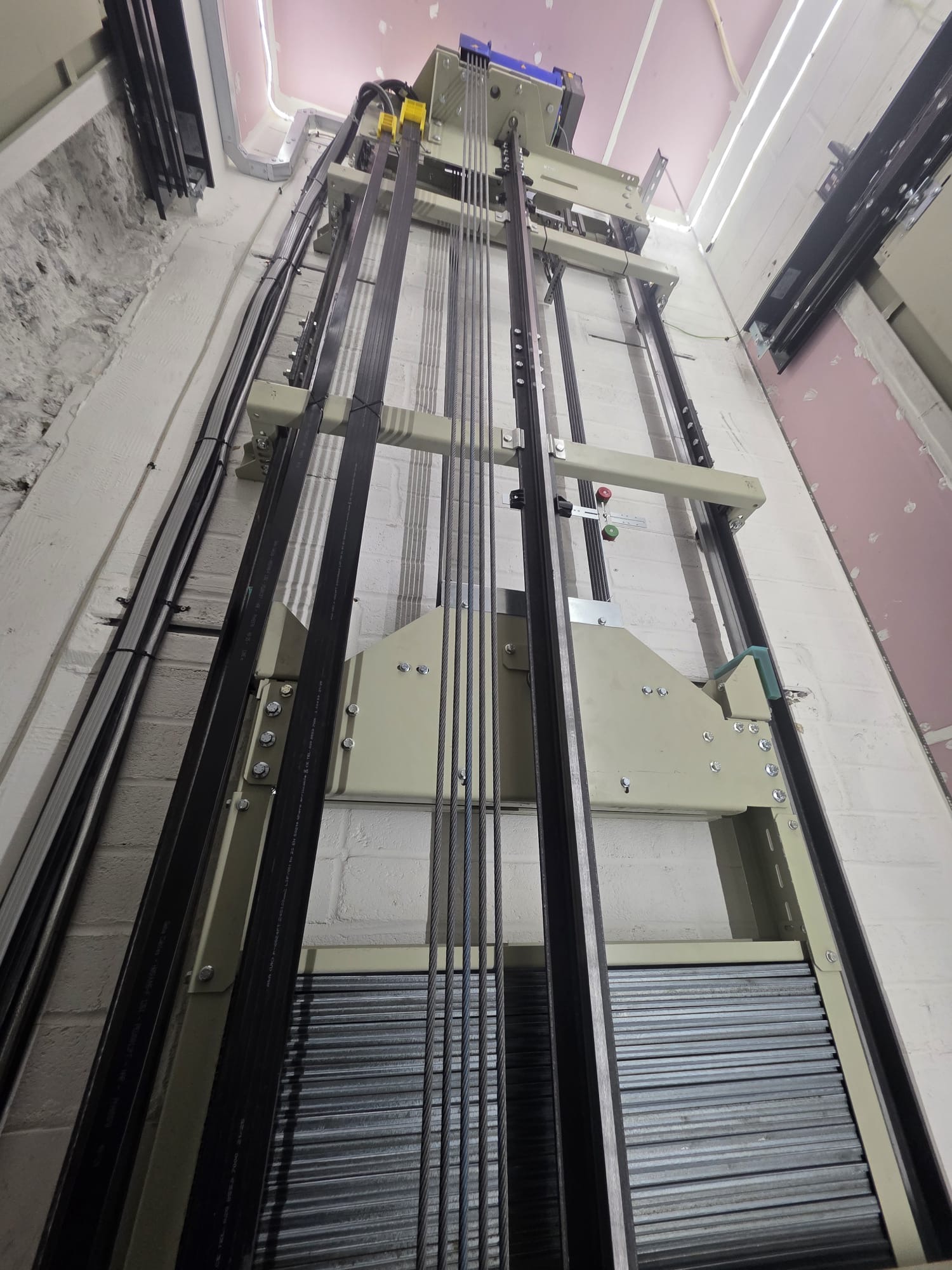 Inside view of elevator shaft showing metal guide rails, steel cables, bolts, and mechanical components against white and pink walls.