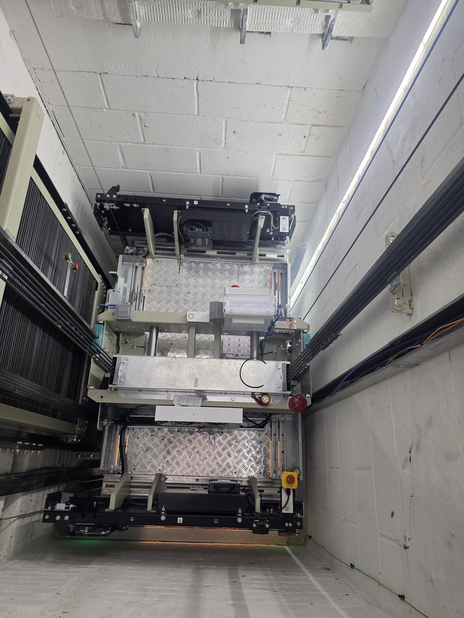 View looking up inside an elevator shaft showing the top section of an elevator car with metal framework, cables, pulleys, safety devices, and control mechanisms mounted on walls with white bricks.
