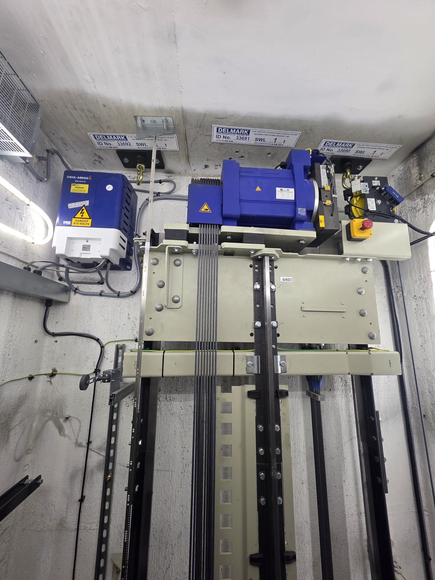 Upper part of an elevator motor system installed in a small room with white textured walls, featuring a blue motor unit connected to multiple steel cables running vertically, safety labels, and electrical components mounted on the ceiling and walls.