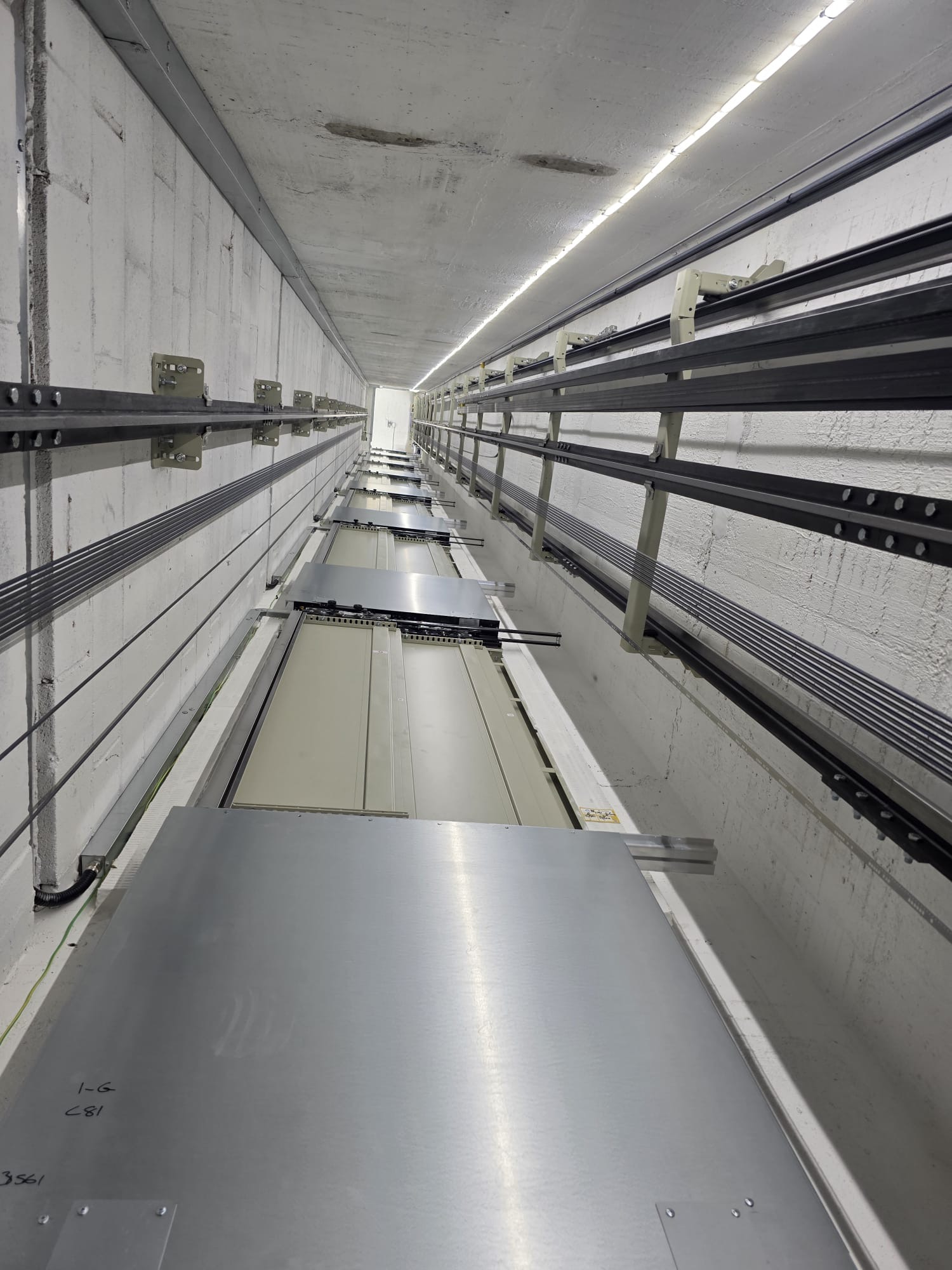 View looking up inside an elevator shaft showing steel cables, guide rails, and elevator doors with a small lighted opening at the top.