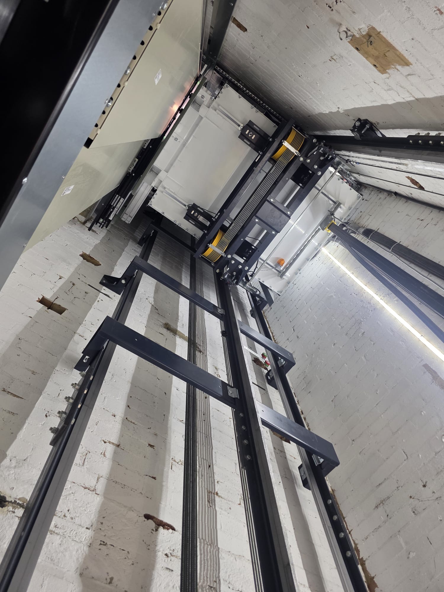View looking up inside an elevator shaft showing black steel rails and cables against white brick walls with the elevator car at the top.
