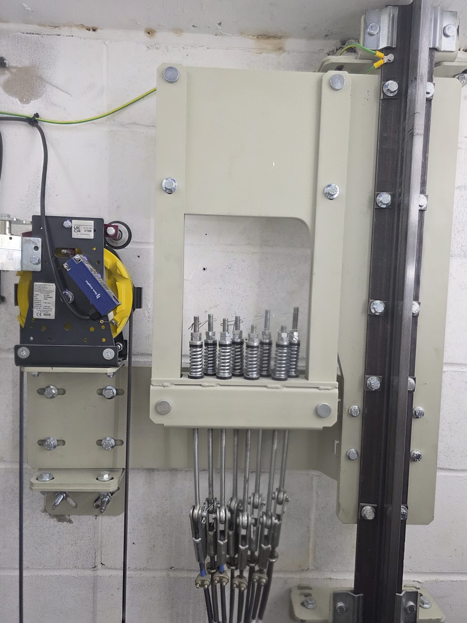 Close-up of mechanical assembly mounted on a wall with seven tension springs each connected to cables with turnbuckles and a yellow and black component attached to the left side.