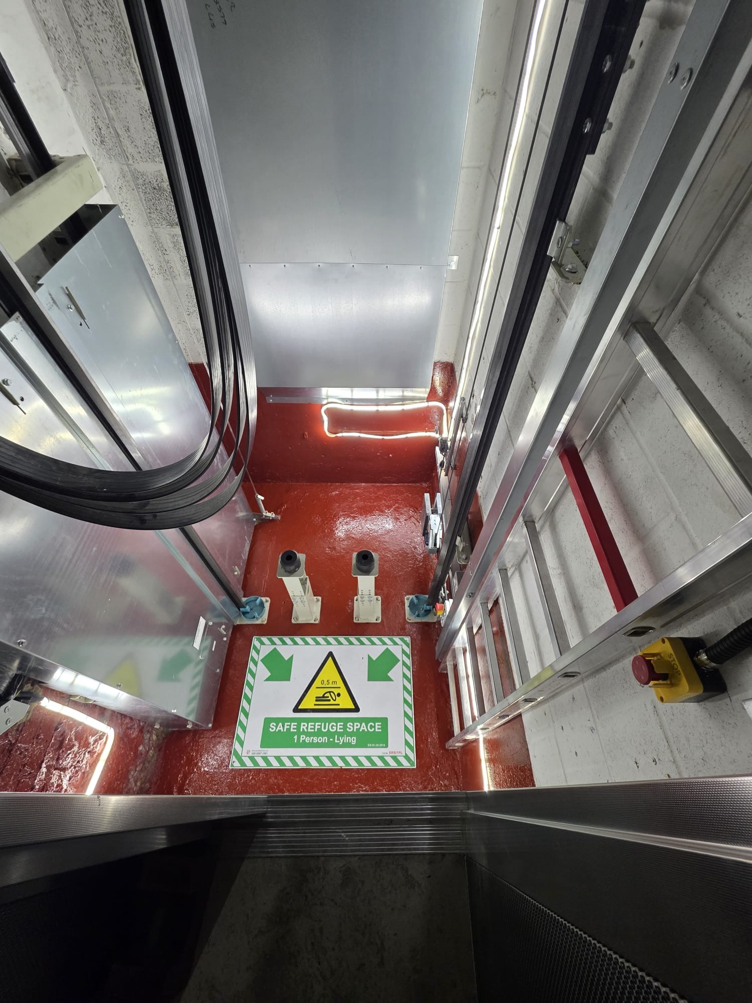 Inside an industrial elevator shaft showing metal rails, cables, a red floor, and a green and white sign reading 'SAFE REFUGE SPACE 1 Person - Lying' with a warning symbol indicating 0.5 meters space.