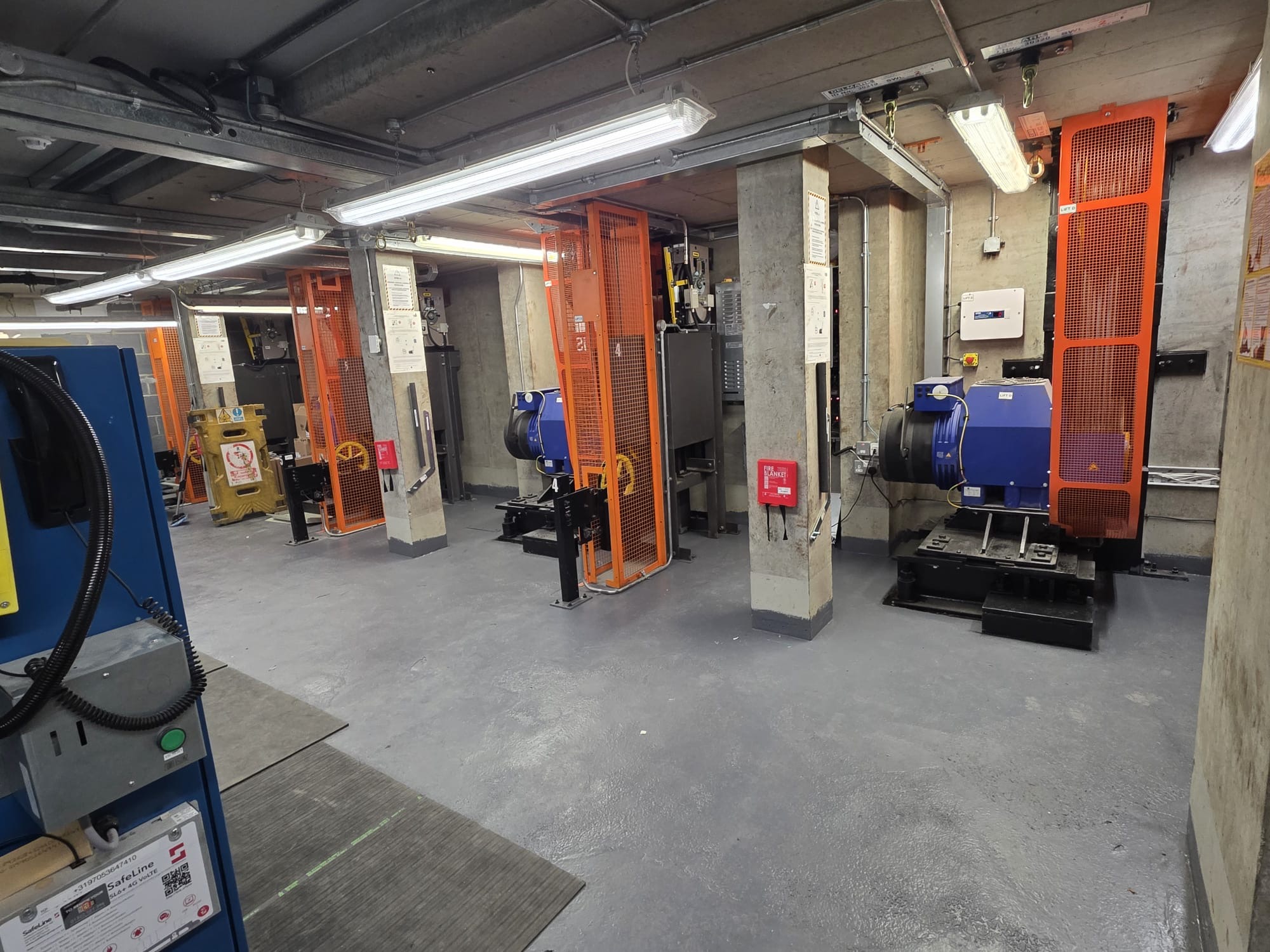 Industrial room with concrete columns and floor, housing several large mechanical machines with blue and orange parts enclosed by orange safety cages under fluorescent ceiling lights.