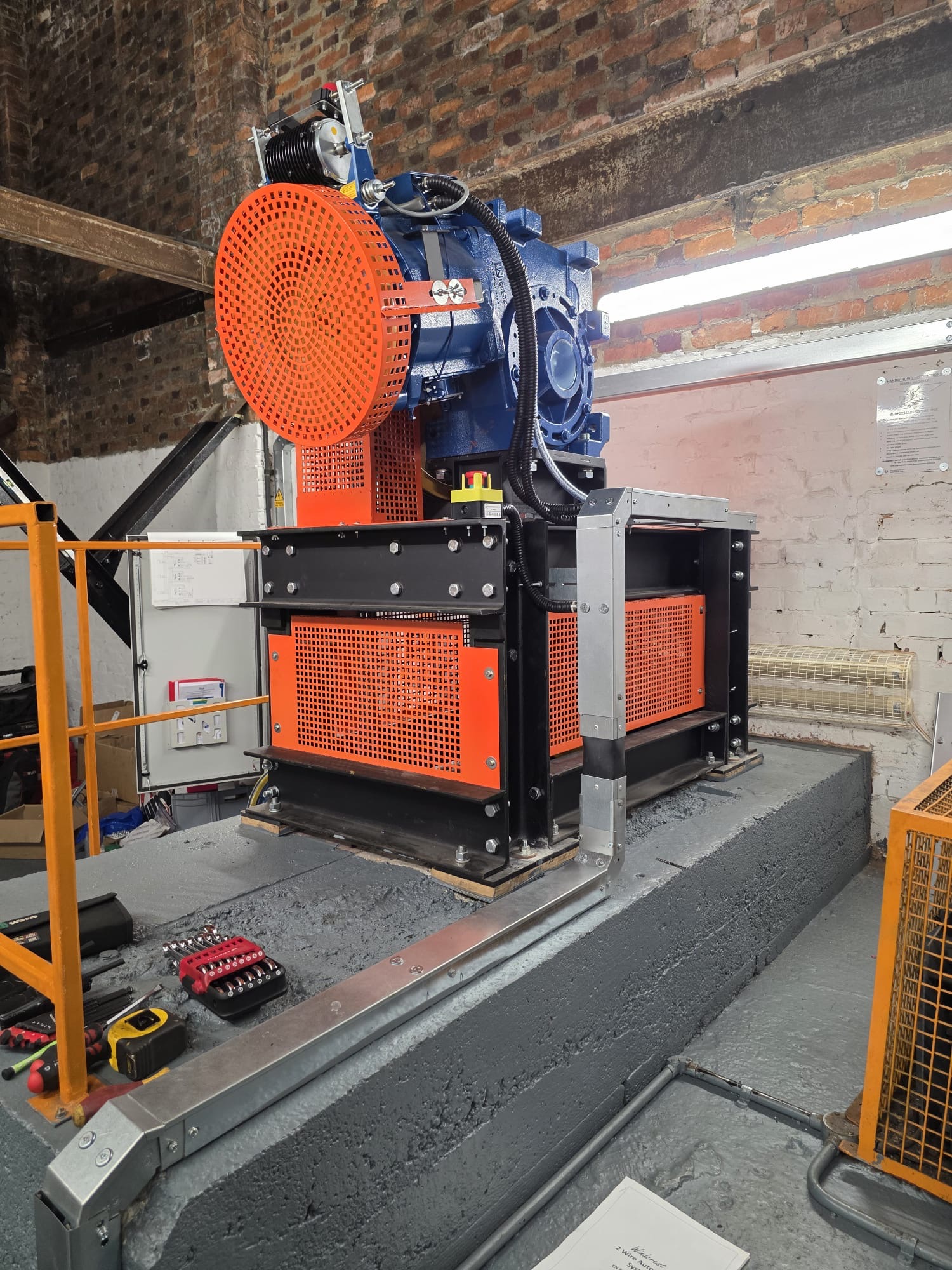 Industrial machine with orange protective grilles and blue motor mounted on a gray concrete platform inside a brick-walled workshop with tools and electrical components nearby.