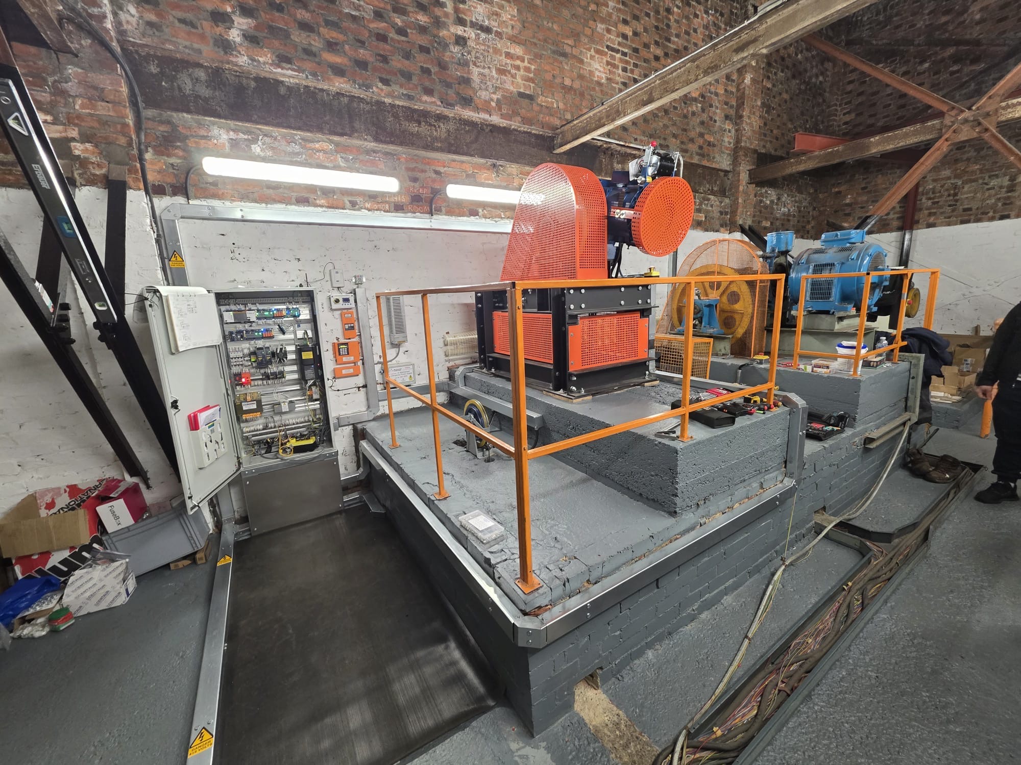 Industrial room with machinery including an orange-covered motor and blue equipment on raised platforms surrounded by orange railings, an open electrical panel on the left, cables running along the floor, and a person partially visible on the right.