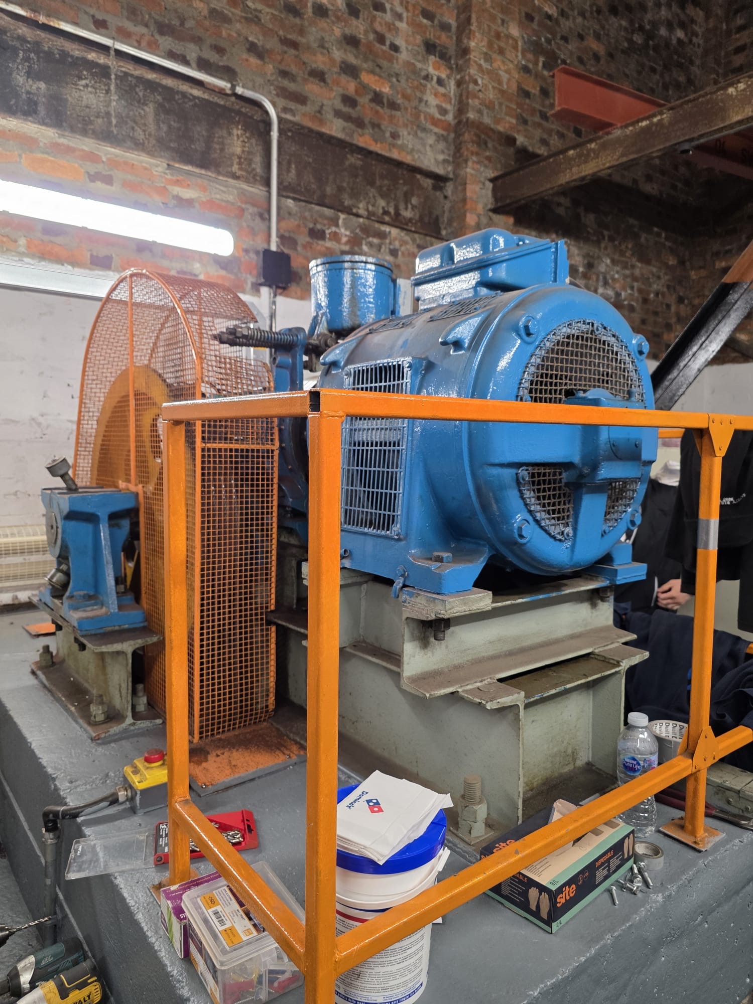 Blue industrial electric motor mounted on a grey base inside a brick building, surrounded by orange safety railing with various tools, boxes, and a water bottle near the base.