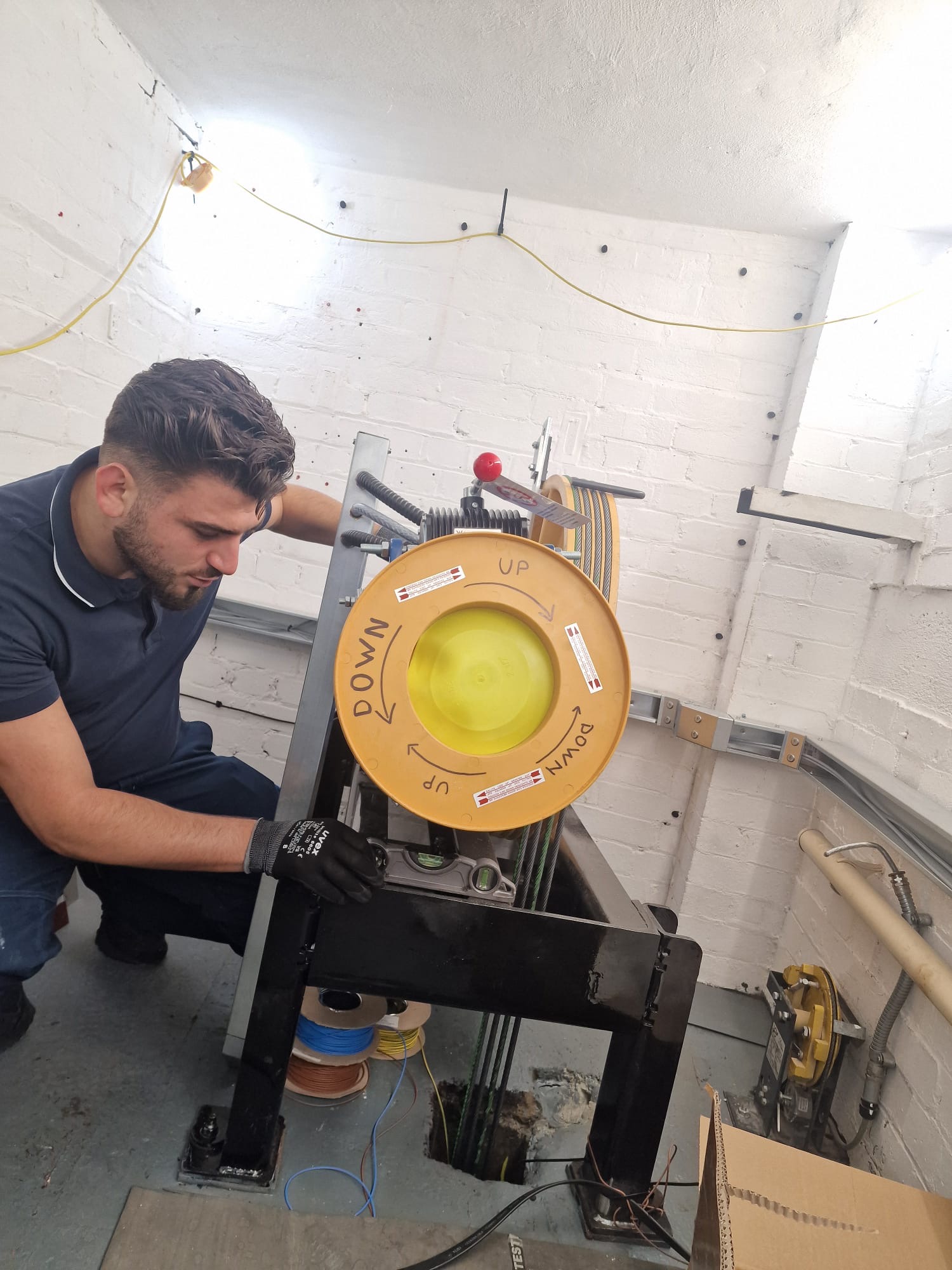 A man wearing a navy blue polo and black gloves is using a level tool to check a large machine with yellow wheels labeled with directional arrows and 'UP' and 'DOWN' in a white-walled room.