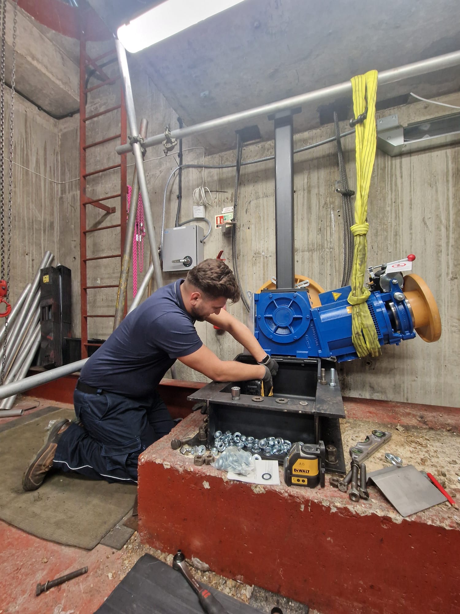 A man wearing a navy blue shirt, gloves, and work pants kneels on the floor assembling or repairing a large blue industrial machine secured with yellow straps in a concrete-walled workshop. Various bolts, nuts, tools, and parts are scattered on a red concrete platform in front of him. In the background, there is a red ladder, metal pipes, and electrical wiring mounted on the wall.