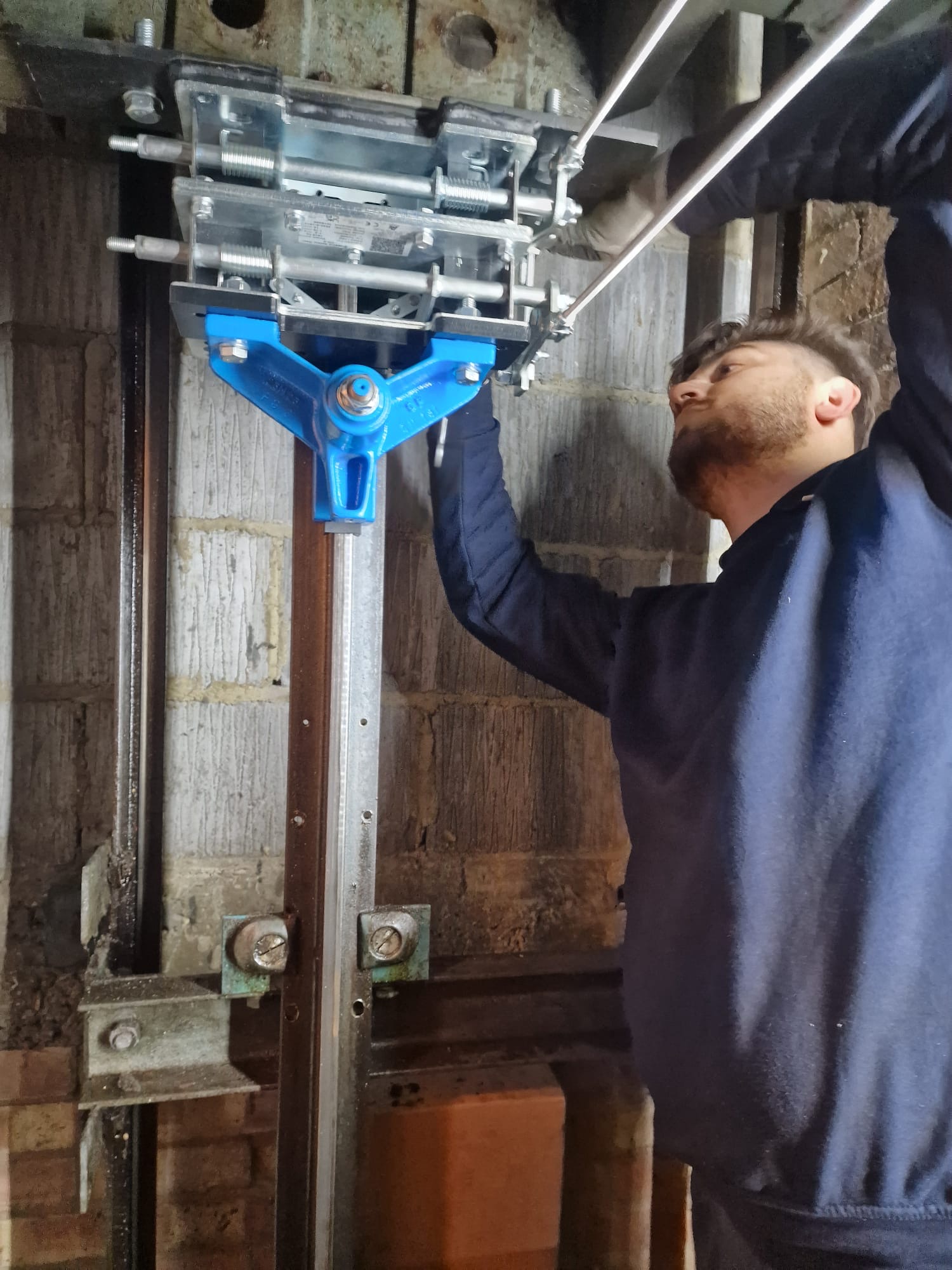 Man with light beard wearing a dark sweatshirt is adjusting or installing a mechanical component with a blue triangular part mounted on vertical metal rails against a wall made of concrete blocks.